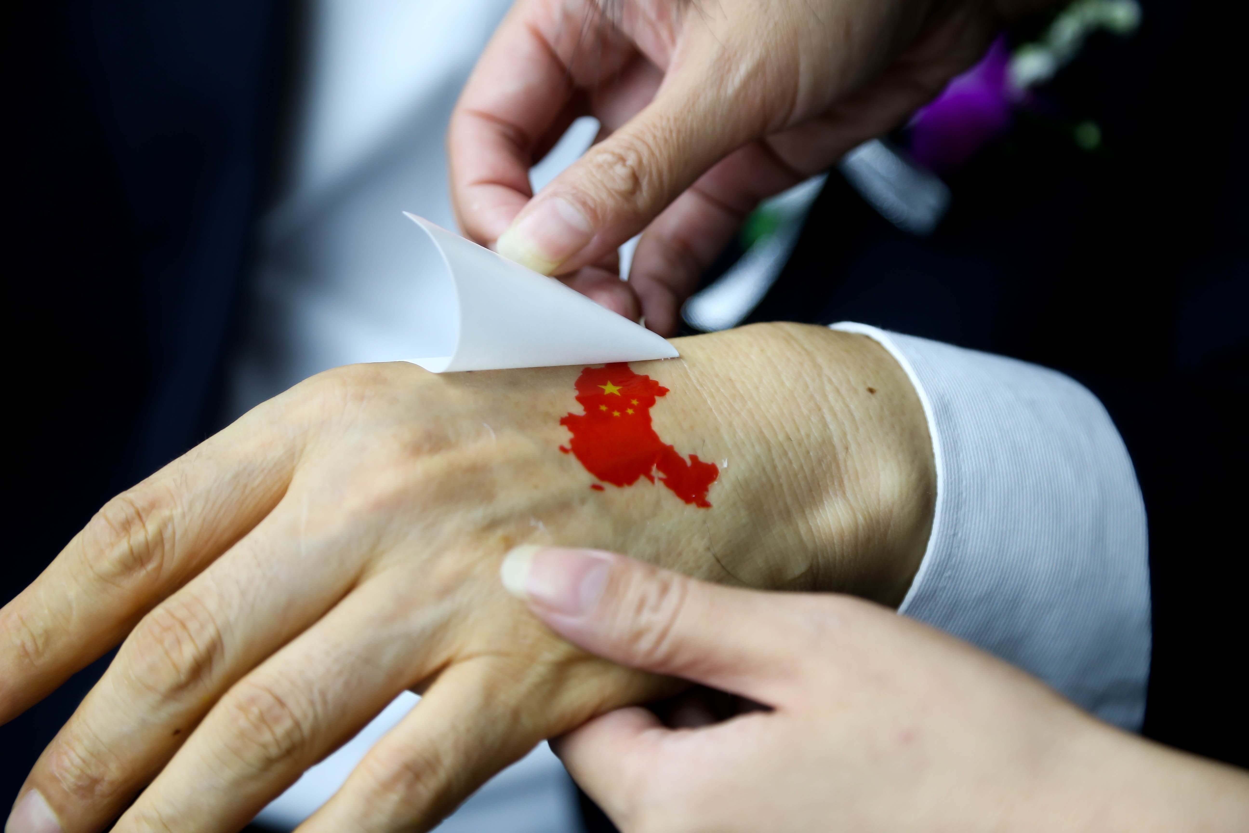 活动现场工作人员正为参会伙伴在手背粘贴融合中国轮廓与五星红旗元素的爱国主题贴纸，动作细致，氛围庄重热忱，充分展现安利伙伴的家国认同感与活动正向主旋律。
