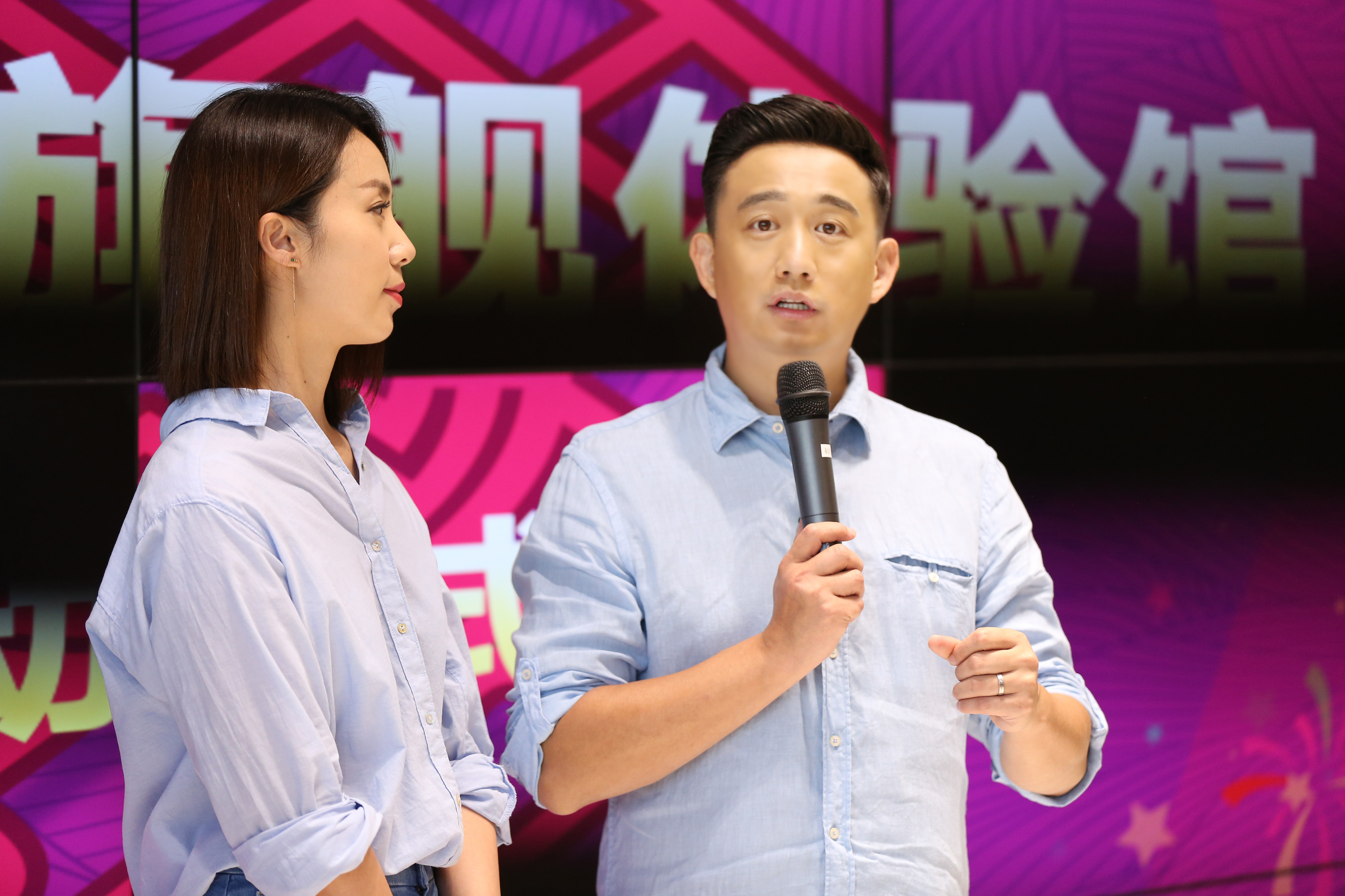 背景大屏印有“体验馆”核心字样，手持麦克风的男士正在现场进行分享，身旁女士侧耳倾听，二人着装简约清爽，现场氛围轻松专业，充分展现安利品牌活动活力务实的特质，传递正向积极的品牌理念。