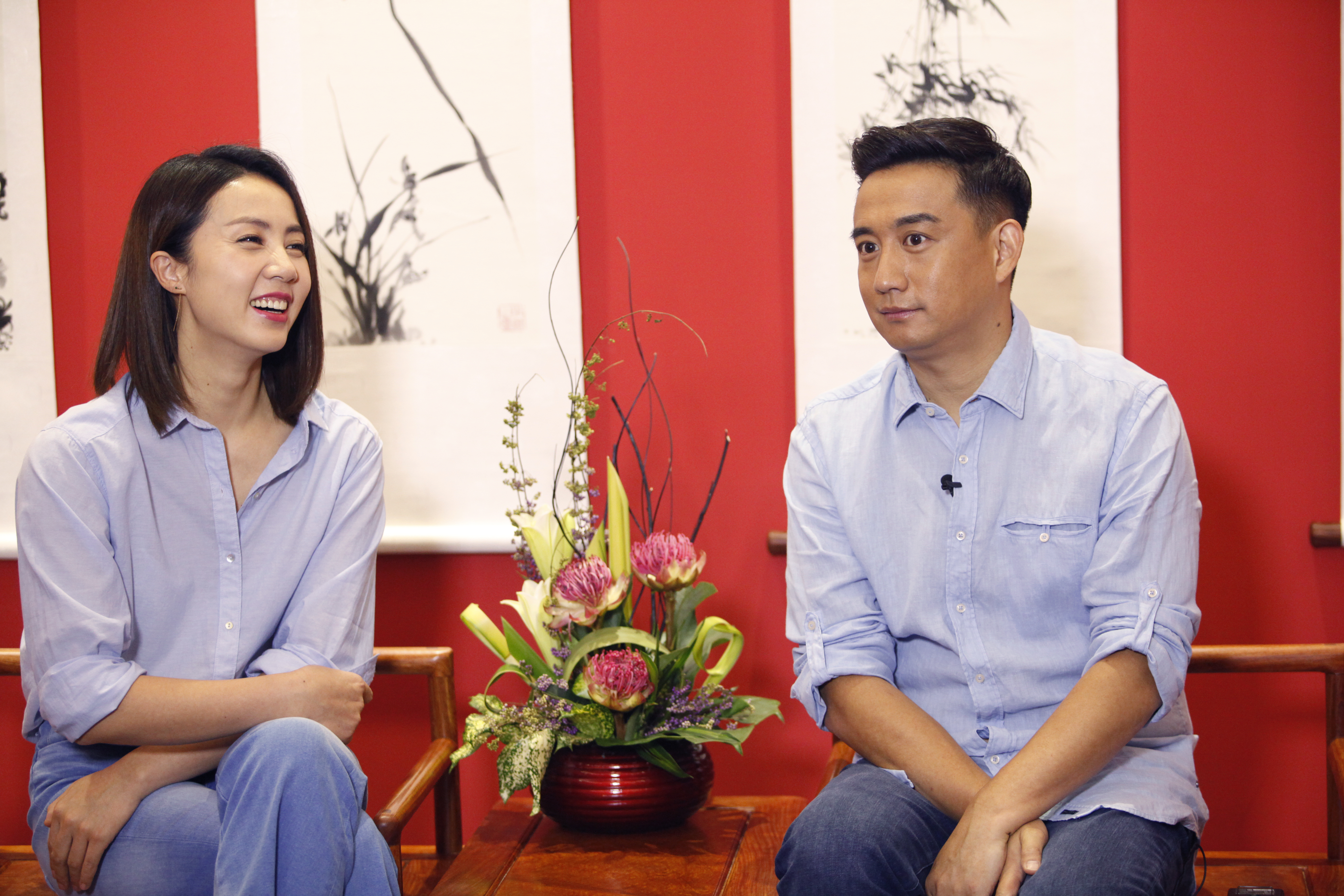 本次安利品牌活动访谈环节现场，黄磊、孙莉两位嘉宾身着浅蓝衬衫相对落座，中间桌台摆放精美花艺陈设，背景红墙搭配水墨国画，整体环境雅致大方，现场氛围轻松融洽，二人正投入参与交流。