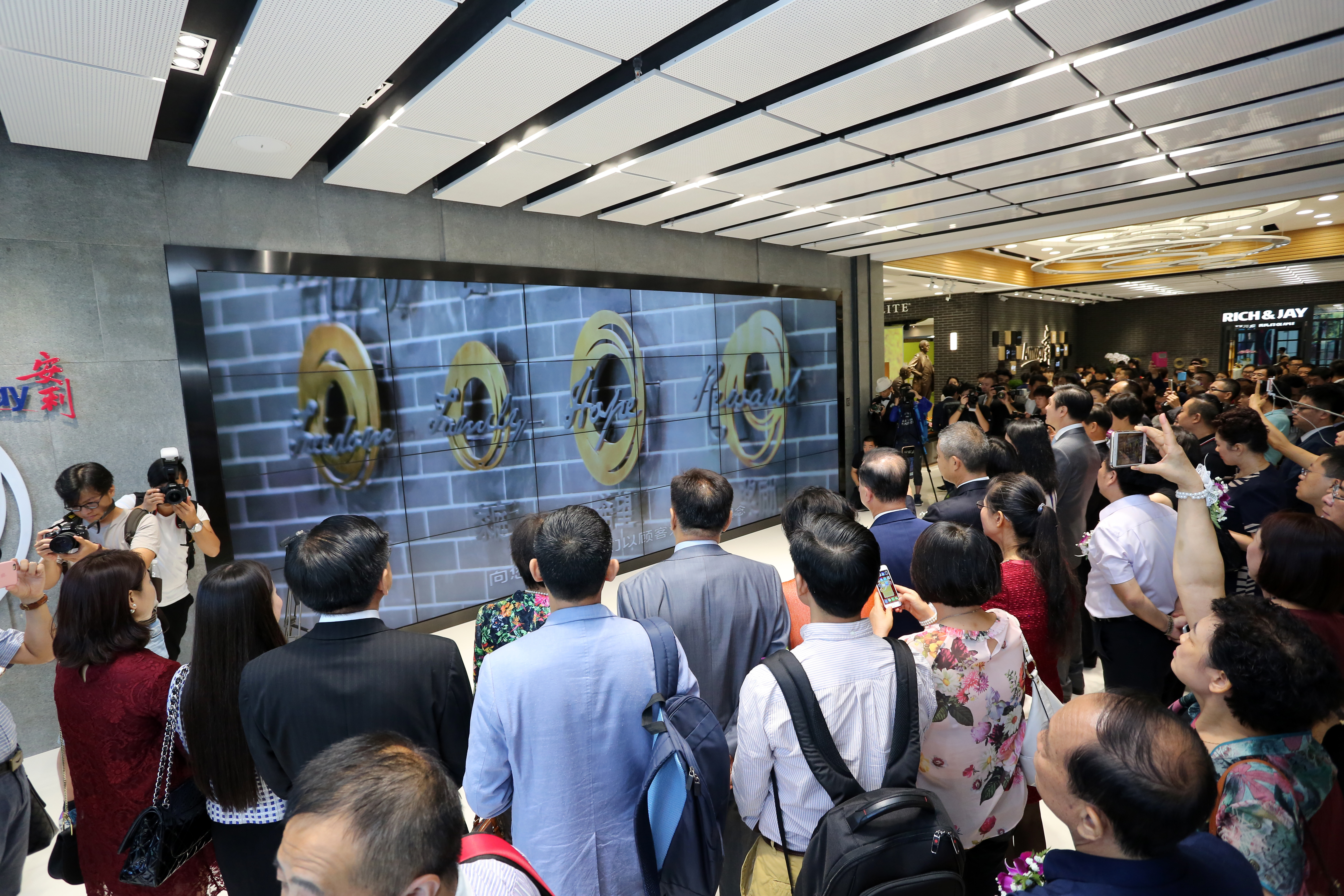 现场墙面设有安利品牌标识，众多嘉宾、媒体工作者齐聚场地，共同观看前方大型拼接展示屏，屏上呈现“自由、家庭、希望、奖励”的品牌核心理念内容，氛围热烈庄重，不少人员举设备拍照记录，尽显对活动的高度关注。