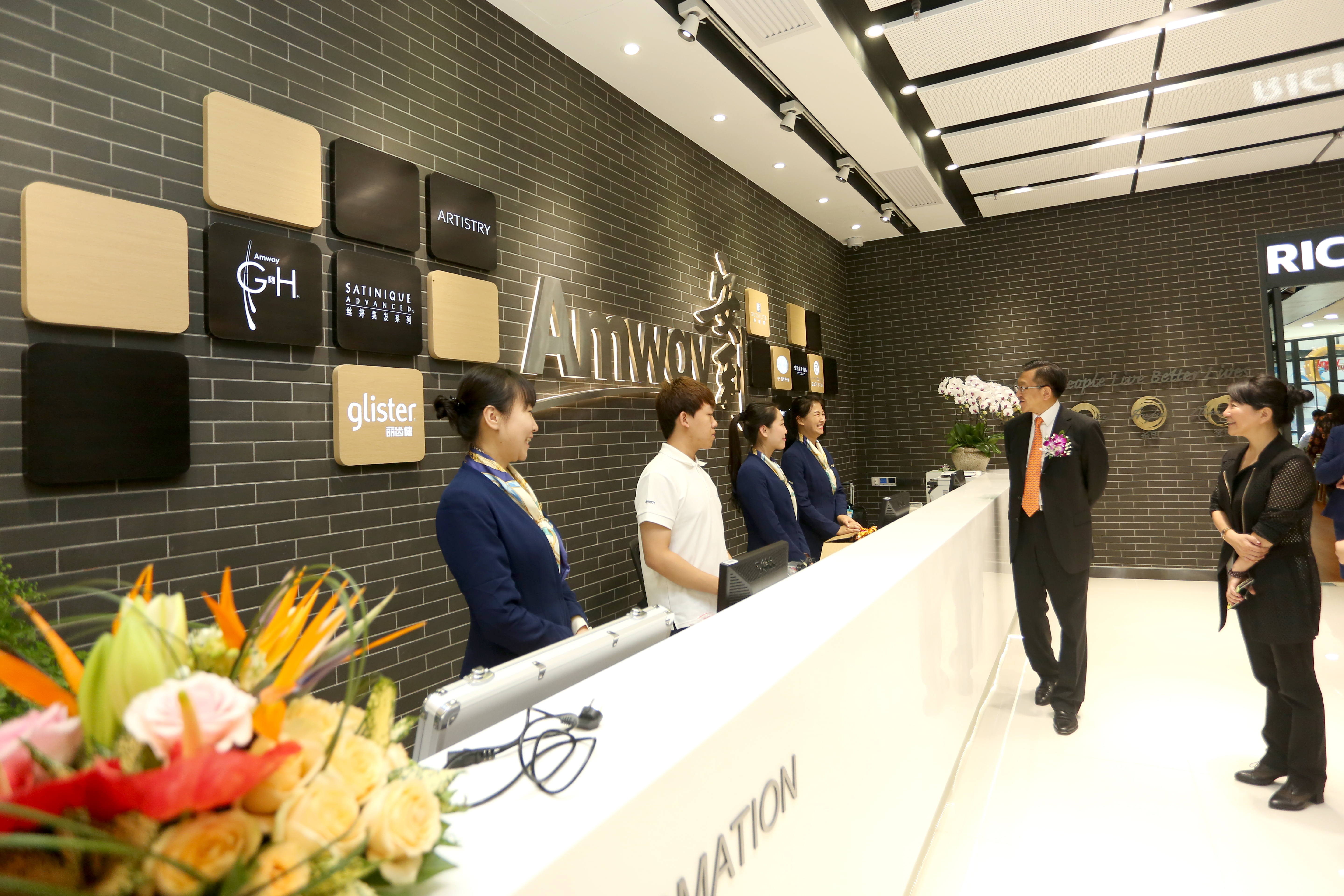 画面背景墙展示有Amway标识及雅姿ARTISTRY、丽齿健glister、丝婷SATINIQUE、Amway GH等旗下品牌标识，数名工作人员整齐值守前台，正与到访来宾友好交流，空间明亮整洁，氛围融洽专业，尽显安利线下场所的优质服务风貌。