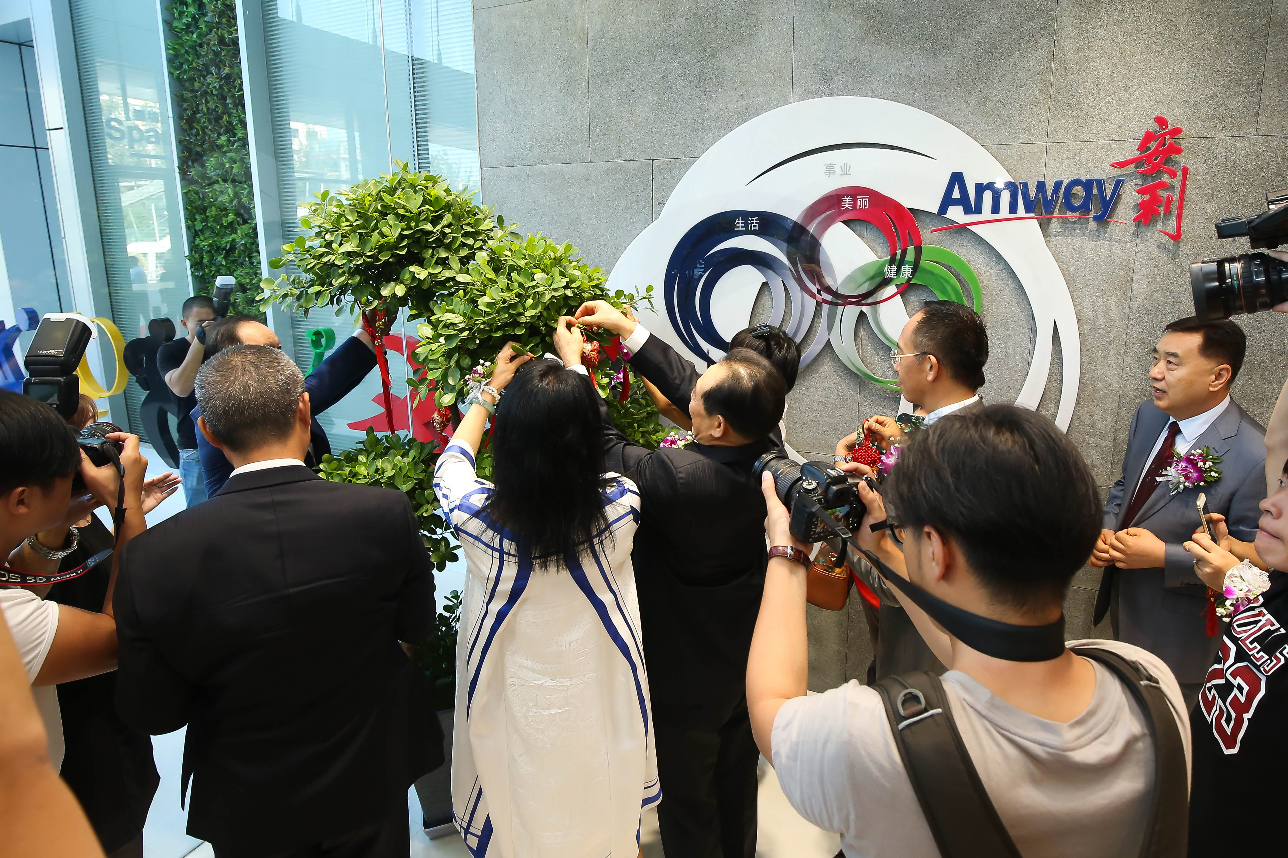 背景墙面印有“Amway”“安利”字样及标注“生活、事业、美丽、健康”的品牌标识，多位嘉宾正携手为绿植悬挂祈福饰品，现场有多名媒体工作人员持摄影设备记录盛况，氛围热烈喜庆，尽显安利积极正向的品牌风貌。