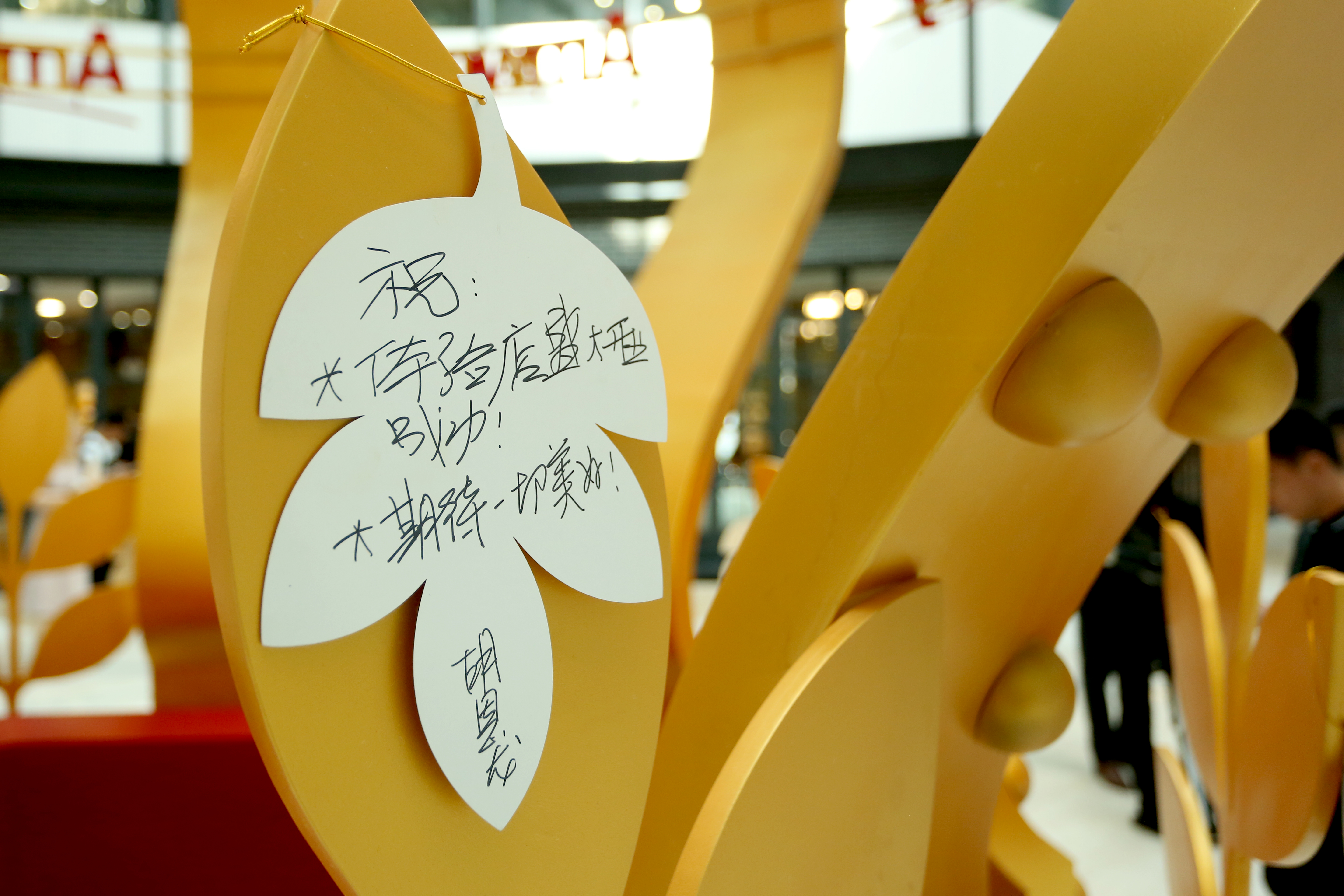 画面展示安利活动现场金色许愿装置上的叶片形手写祝福卡，卡上写有“祝：体验店盛大开业成功！期待一切美好！”的祝福语，整体氛围温暖诚挚，承载着参与者对门店开业的美好期许，尽显活动正向暖心的氛围。