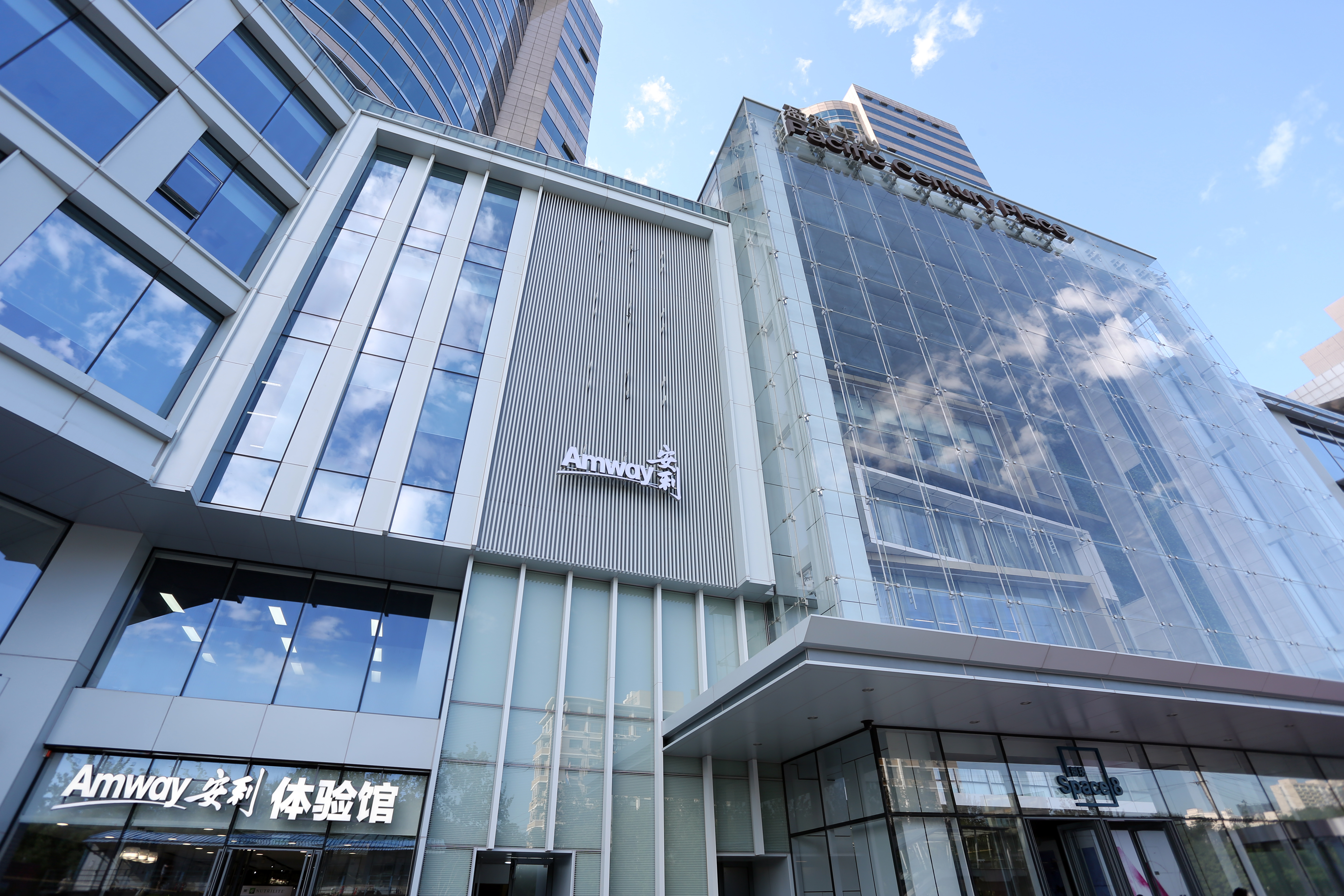 晴朗蓝天之下，现代化商业楼宇整洁大气，建筑墙面醒目设置“Amway安利”标识，底层入口处标注“Amway安利 体验馆”字样，玻璃幕墙倒映云影，整体场景明亮开阔，彰显安利品牌专业正向的品牌质感。