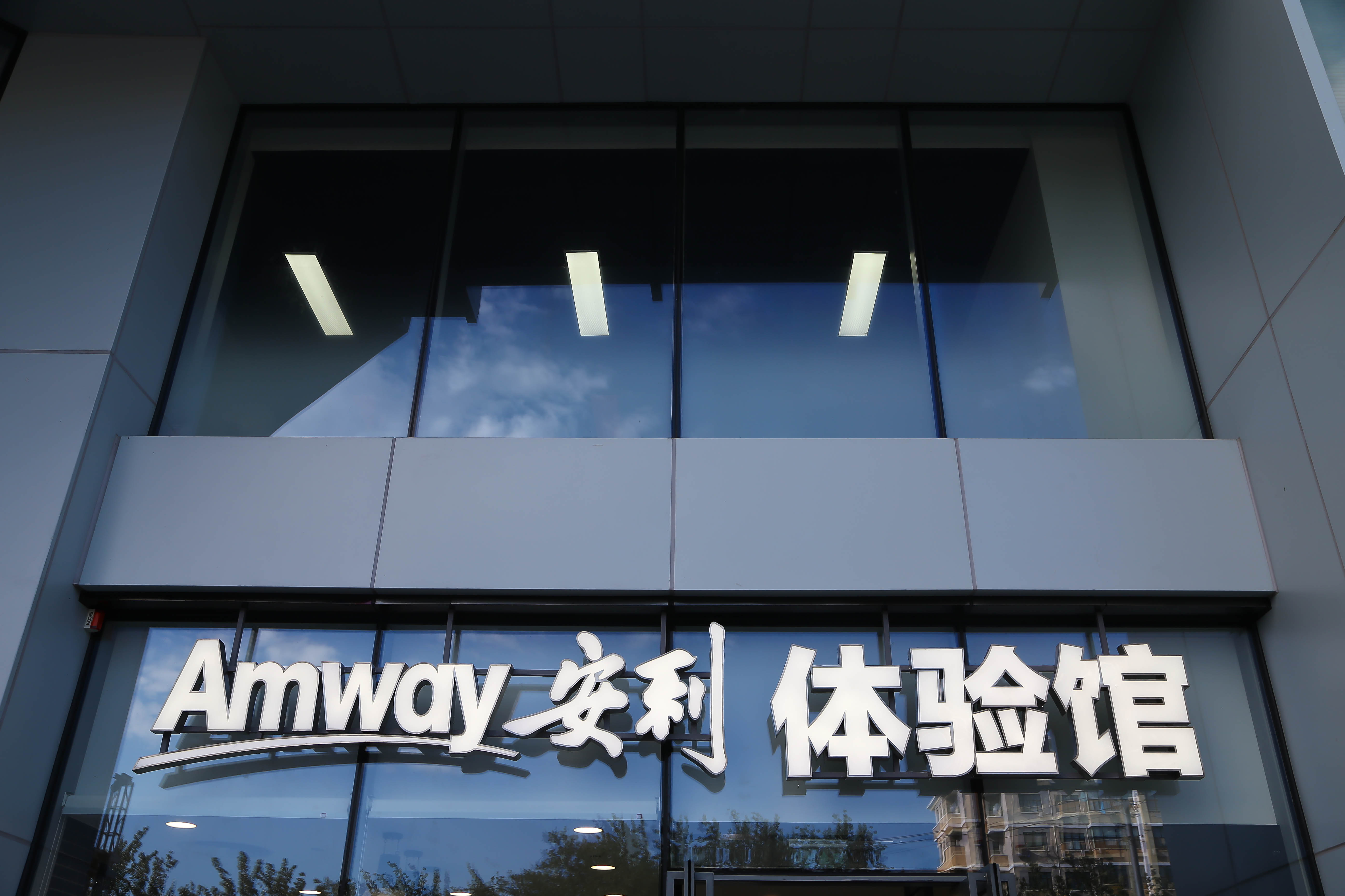 画面呈现安利体验馆门头场景，醒目的“Amway 安利 体验馆”立体标识清晰可见。建筑采用简约现代设计风格，玻璃幕墙澄澈透亮，反射着蓝天白云与周边景致，整体氛围大气专业，尽显安利线下体验场景的品质感与品牌调性。