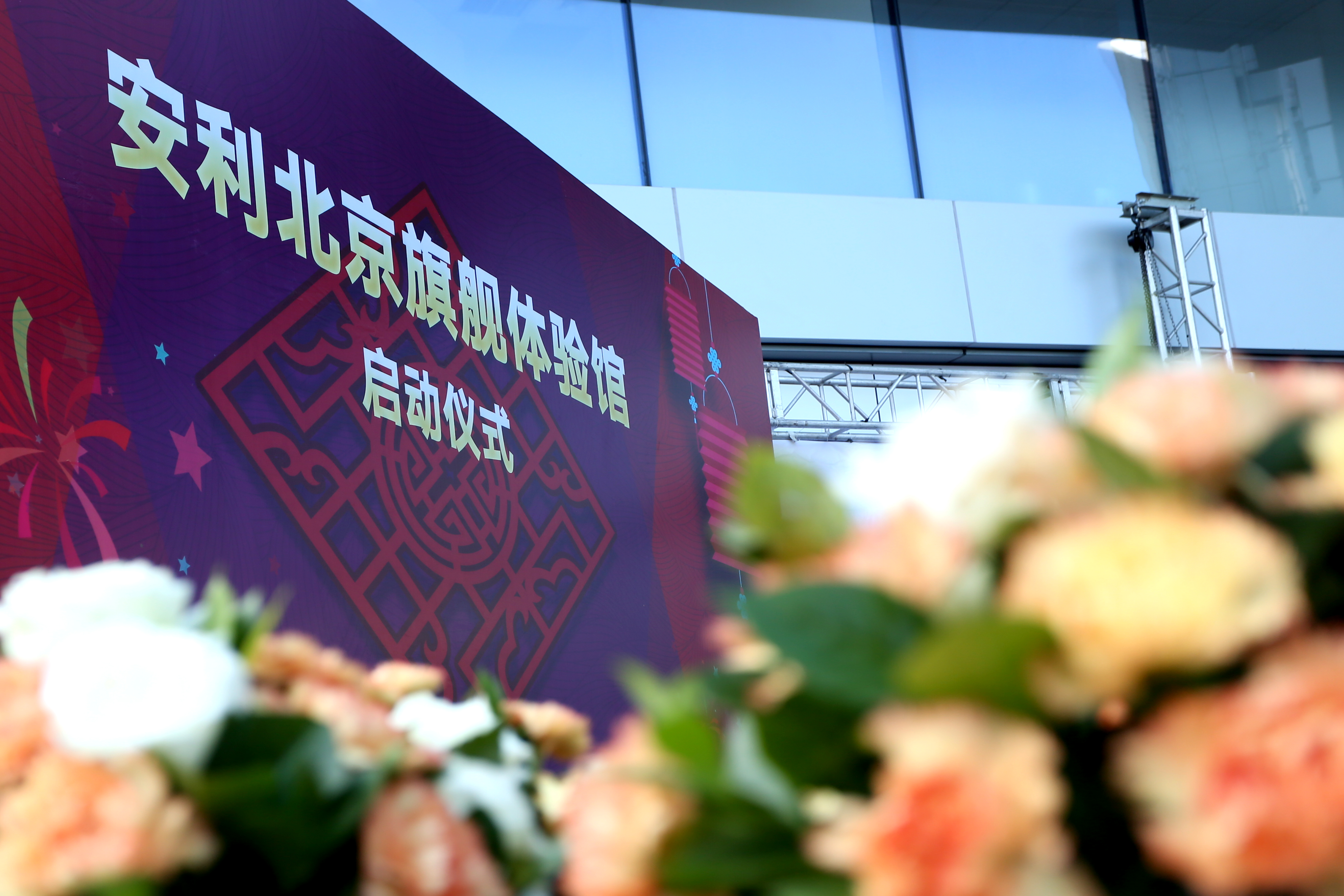 画面为安利北京旗舰体验馆启动仪式现场，印有活动主题的紫色背景板搭配中式吉祥纹样，彰显喜庆氛围。前景错落的花艺柔和雅致，背景可见场馆玻璃幕墙与活动搭建桁架，整体场景隆重正式，凸显品牌对本次活动的重视。