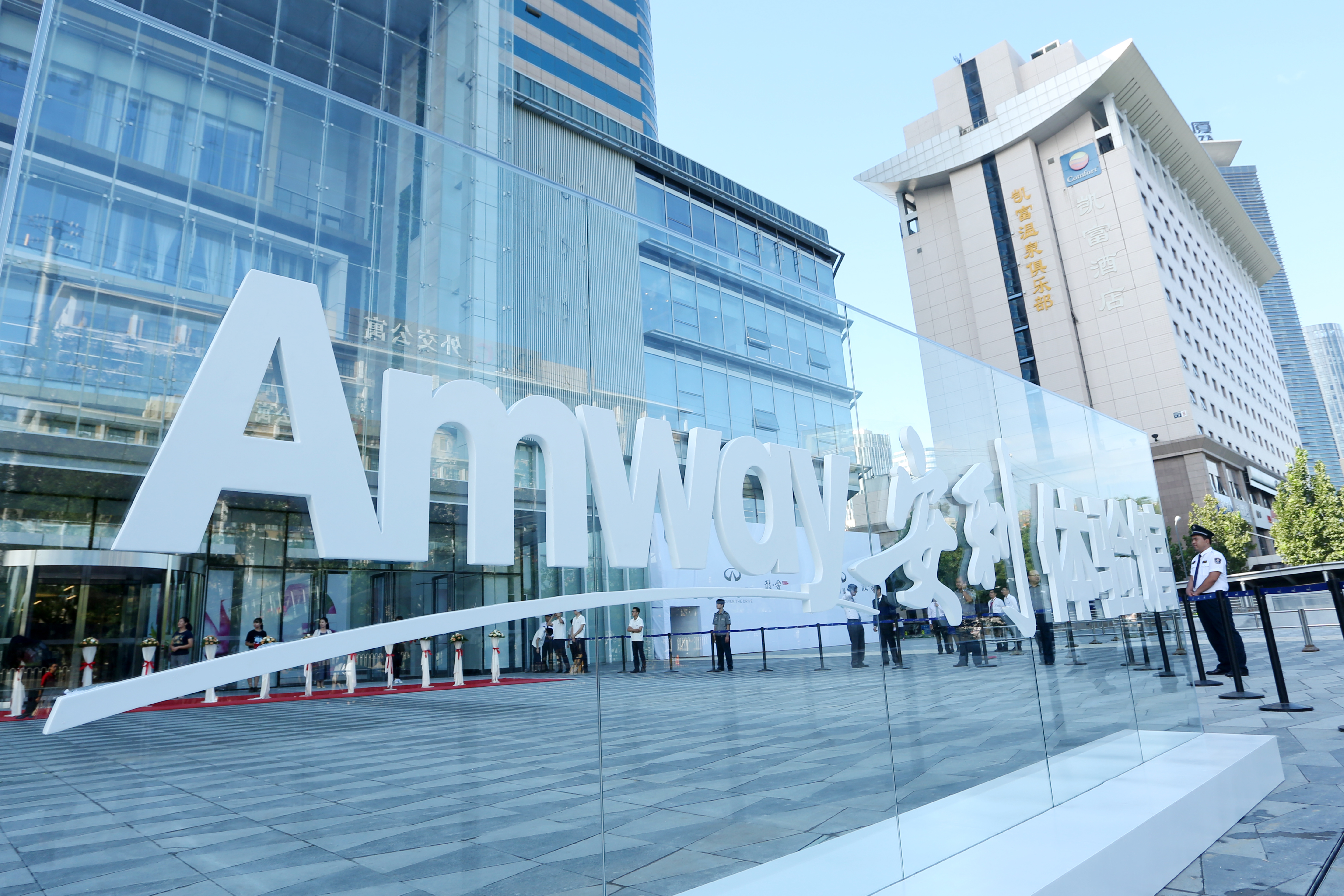 晴朗天气下，安利体验馆外设有大型立体“Amway”及“安利体验馆”品牌标识，背景为现代化玻璃幕墙场馆与城市商务楼宇，现场场地开阔整洁，工作人员有序值守，整体氛围正式大气，彰显安利品牌专业质感。