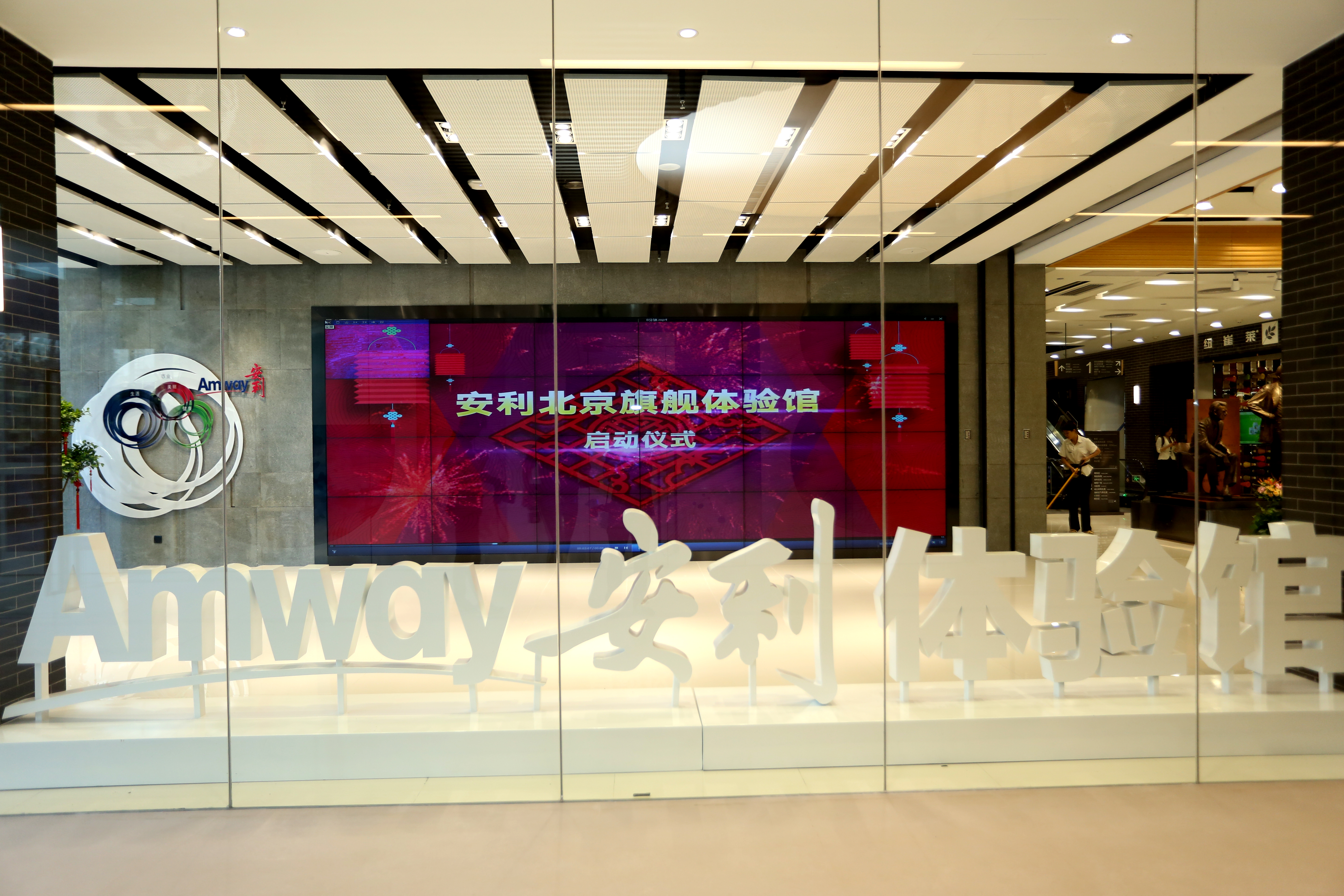画面呈现安利北京旗舰体验馆启动仪式现场，玻璃围挡内设有醒目的“Amway 安利体验馆”白色立体字，背景大屏清晰展示“安利北京旗舰体验馆 启动仪式”字样，墙面配有安利品牌标识，整体空间明亮大气，氛围庄重正式，尽显品牌质感。