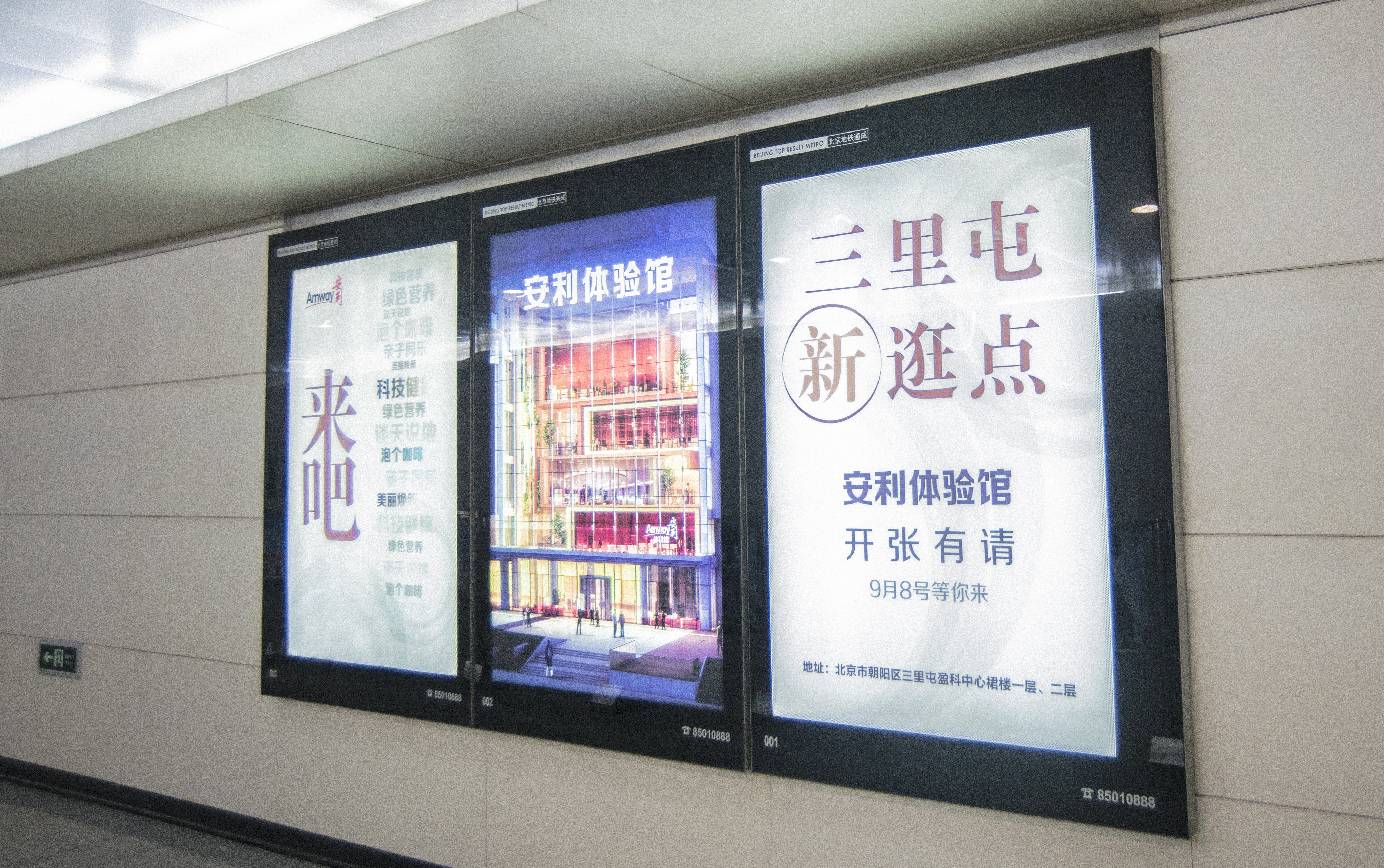 这是北京地铁通道内的三联灯箱宣传广告，左侧广告印有“来吧”“绿色营养”等字样，中间展示安利体验馆外观效果图，右侧标注“三里屯新逛点 安利体验馆开张有请 9月8号等你来”及场馆地址，画面清晰醒目，向公众传递开业邀约信息。