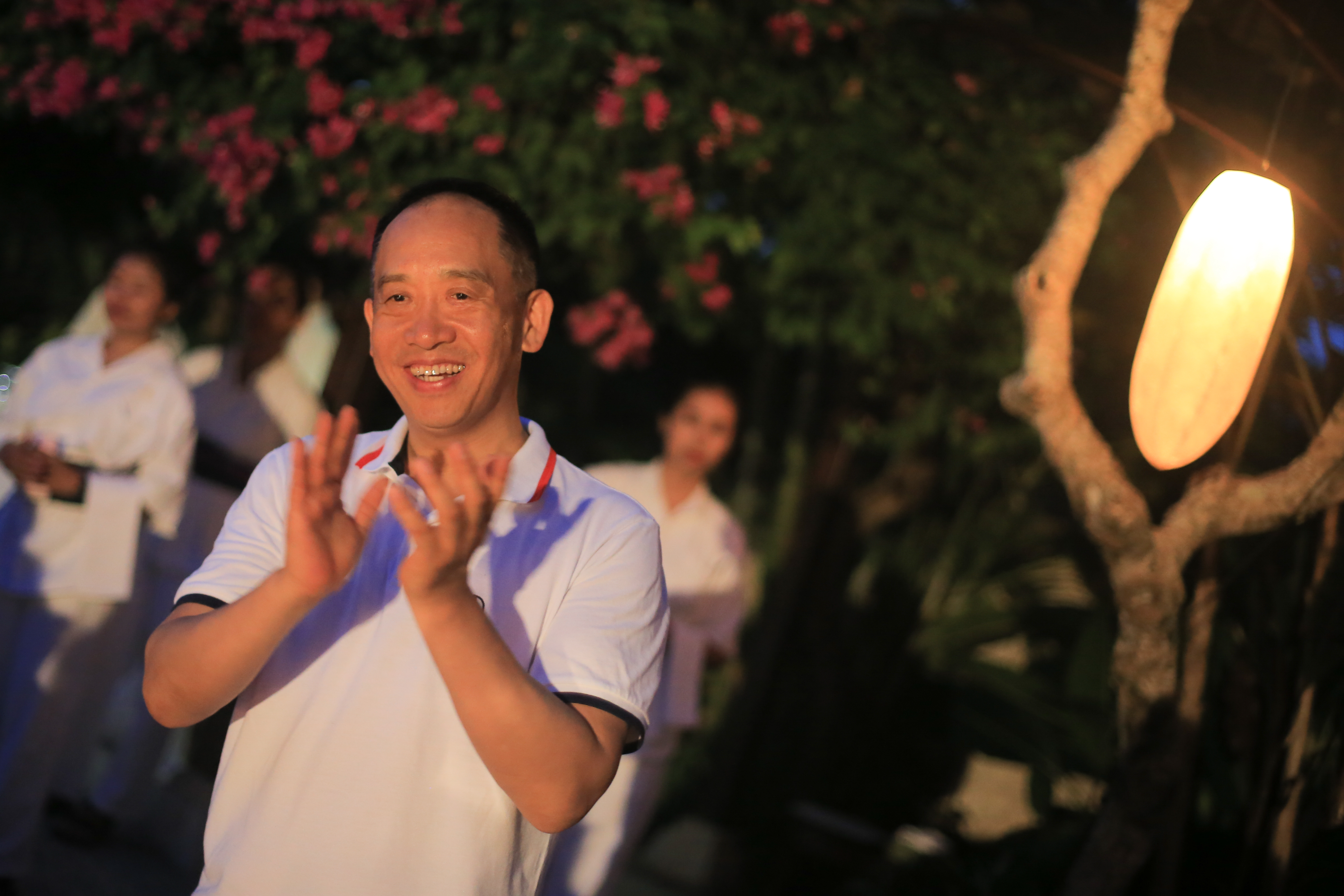 本次安利户外主题活动现场，暖光与花树烘托出温馨松弛的氛围。身着统一白色polo衫的安利领导面带灿烂笑容鼓掌，对现场环节表示认可与赞许，身后其他参会伙伴相伴，整体氛围轻松热烈，尽显团队融洽的积极风貌。