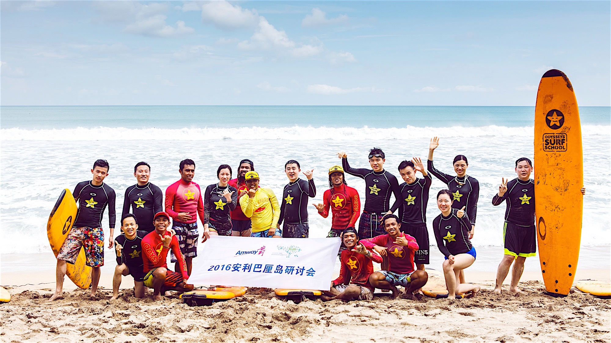 画面为2016安利巴厘岛研讨会冲浪活动现场，碧海白沙的海岸边，参与人员身着冲浪服、搭配冲浪板，围绕印有“2016安利巴厘岛研讨会”的横幅合影，众人笑容灿烂、姿态昂扬，整体氛围活力满满，尽显安利伙伴积极向上的团队风貌。