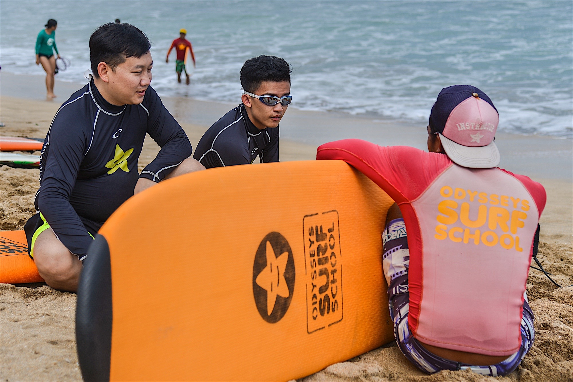 以碧海沙滩为背景，印有“ODYSSEYS SURF SCHOOL”标识的冲浪教练，正与两名身着冲浪服的安利伙伴围坐交流，身旁摆放着同品牌橙色冲浪板，现场氛围轻松融洽，大家认真沟通冲浪技巧，尽显安利团队户外活动的活力与向上风貌。