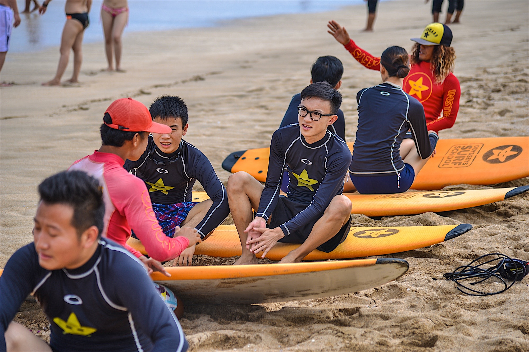 活动在沙滩开展，冲浪板印有“OUTERSETS SURF SCHOOL”标识，安利伙伴们身着专业冲浪服围坐，在教练指导下学习冲浪相关知识，现场氛围轻松活跃，充分展现出安利伙伴朝气蓬勃、乐于挑战、积极交流的良好精神风貌。
