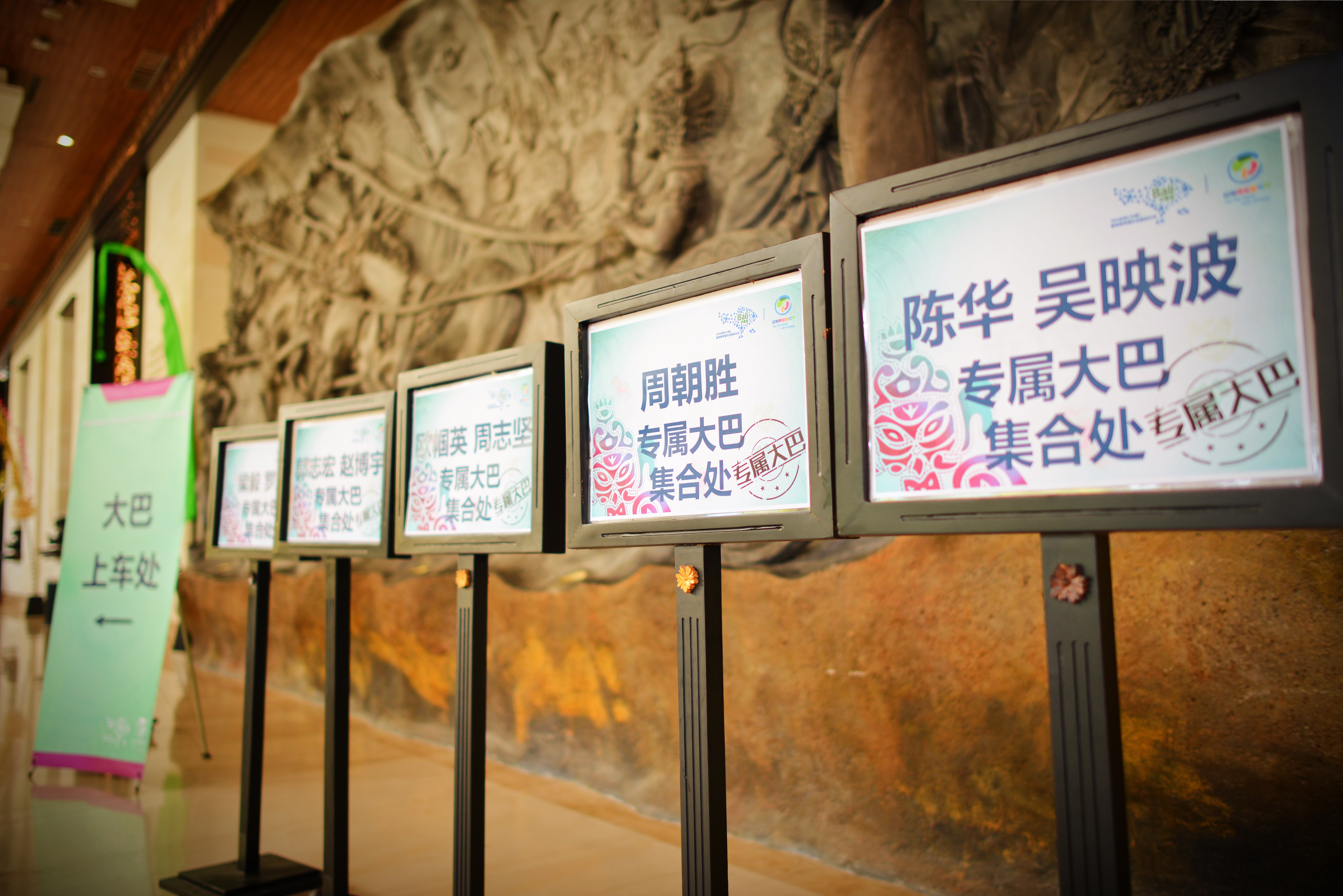 画面展示活动场馆内的乘车指引区域，整齐立有多块标注不同姓名的“专属大巴集合处”标识牌，旁侧还有指向清晰的“大巴上车处”指引展架，背景是艺术浮雕墙，整体布置清晰有序，尽显活动筹备的细致贴心，为参会人员提供明确的乘车引导。