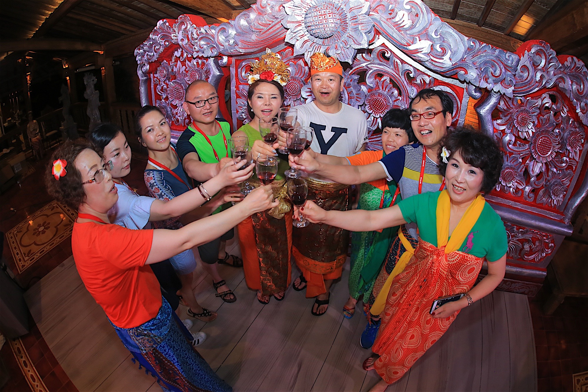 安利异域团建活动现场，多位伙伴身着特色服饰，在精美华丽的异域雕花背景前，共同高举盛有红酒的酒杯碰杯致意，大家面带灿烂笑容，氛围热烈融洽，尽显团队凝聚的喜悦与活动的欢快氛围。