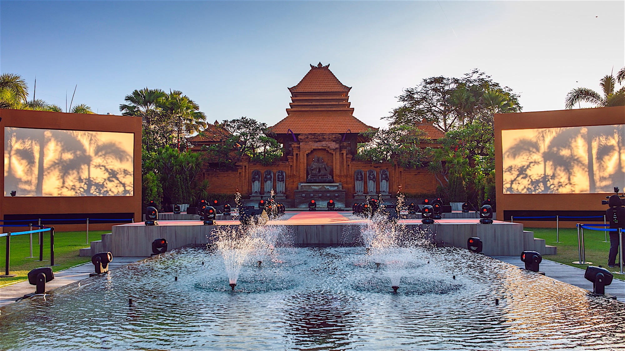 画面为安利户外活动会场，前景灵动喷泉水景鲜活亮眼，中央搭建专业活动舞台，两侧配备高清大屏，背景是东南亚特色传统建筑，热带绿植环绕四周，场地布置规整大气，氛围庄重热烈，尽显活动高端质感。