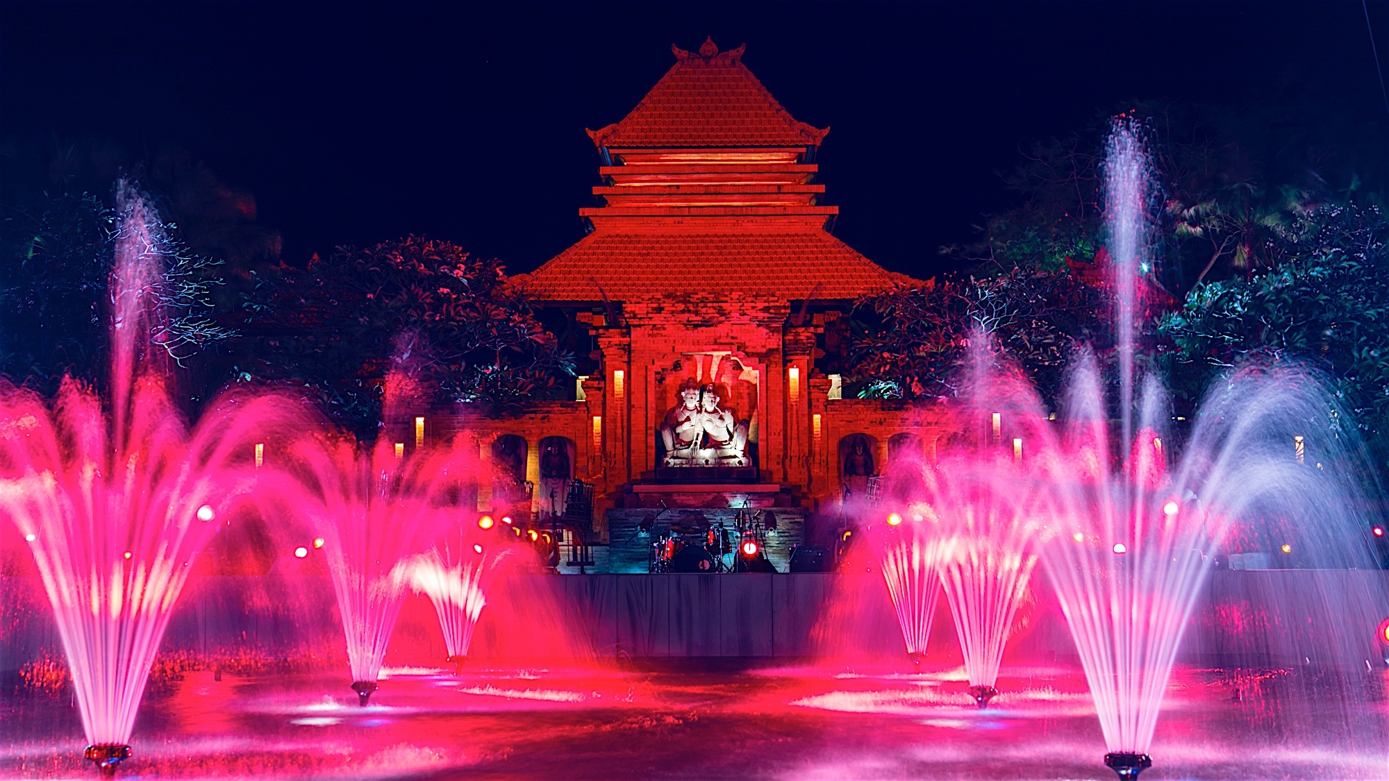 夜幕下，巴厘岛特色传统建筑被暖红灯光点亮，前方彩色光影喷泉灵动喷涌，舞台设施完备，整体氛围绚丽浪漫，是安利为参会伙伴打造的专属特色活动场地，尽显品牌对伙伴的用心礼遇与活动高品质质感。