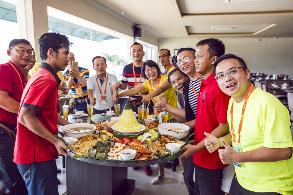 安利团建聚餐现场，佩戴橙色工牌的伙伴们围在摆满特色餐食的圆桌旁，人人脸上洋溢着灿烂笑容，不少伙伴竖起大拇指点赞，现场氛围轻松热烈、融洽温暖，尽显安利团队强大的凝聚力与伙伴们昂扬的归属感。
