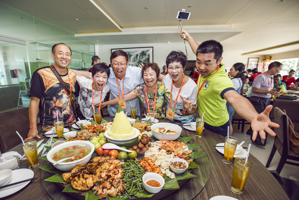 佩戴活动统一挂绳的安利参会伙伴，围在摆满丰盛特色餐食的餐桌旁，众人笑容灿烂情绪高涨，有伙伴举着自拍杆记录欢聚时刻，现场氛围热烈欢快，尽显安利团队融洽的氛围与伙伴们饱满昂扬的精神面貌。