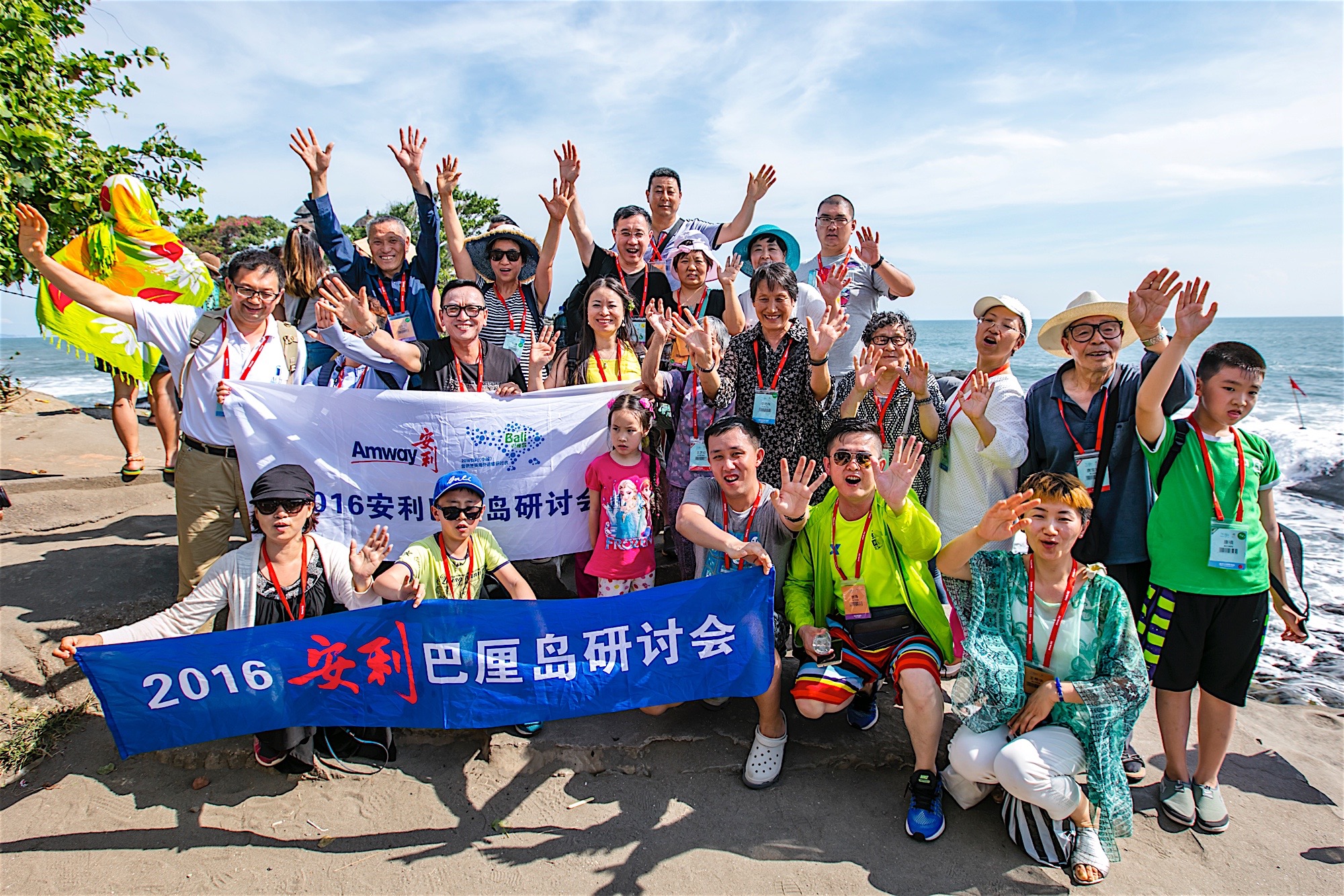 2016安利巴厘岛研讨会举办期间，参会伙伴在海边集体合影。大家手持印有“Amway”“2016安利巴厘岛研讨会”字样的横幅，纷纷面向镜头挥手致意，笑容灿烂，背景是澄澈碧海与晴朗蓝天，氛围轻松欢快，尽显参会伙伴的昂扬状态。