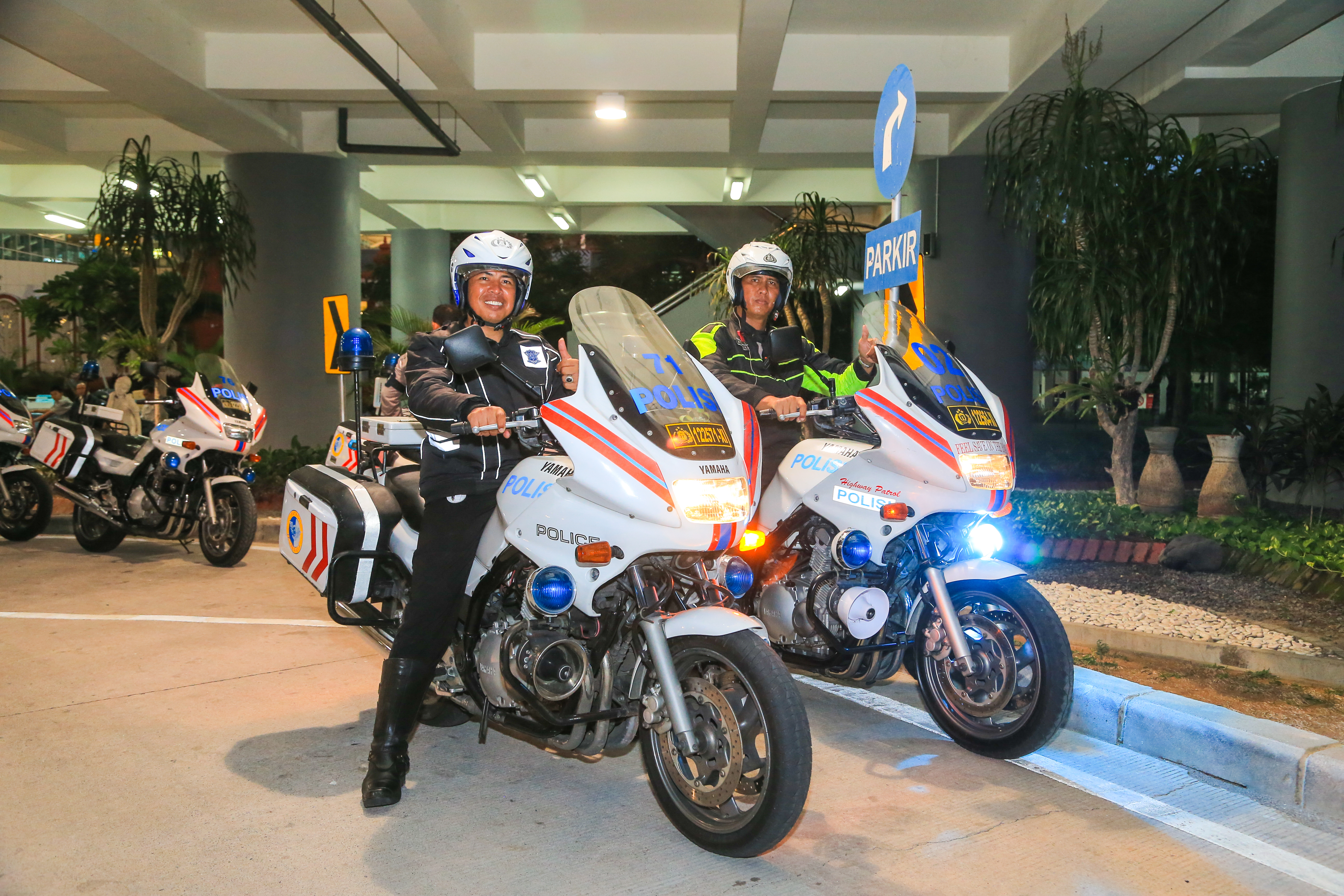 画面中两名印尼警用摩托骑手佩戴头盔，坐在印有“POLIS”字样的雅马哈警用摩托上友好竖起大拇指，周边可见“PARKIR”标识，警用车辆排列整齐，是当地警方为安利相关活动提供出行安全保障，尽显活动高规格与稳妥有序。