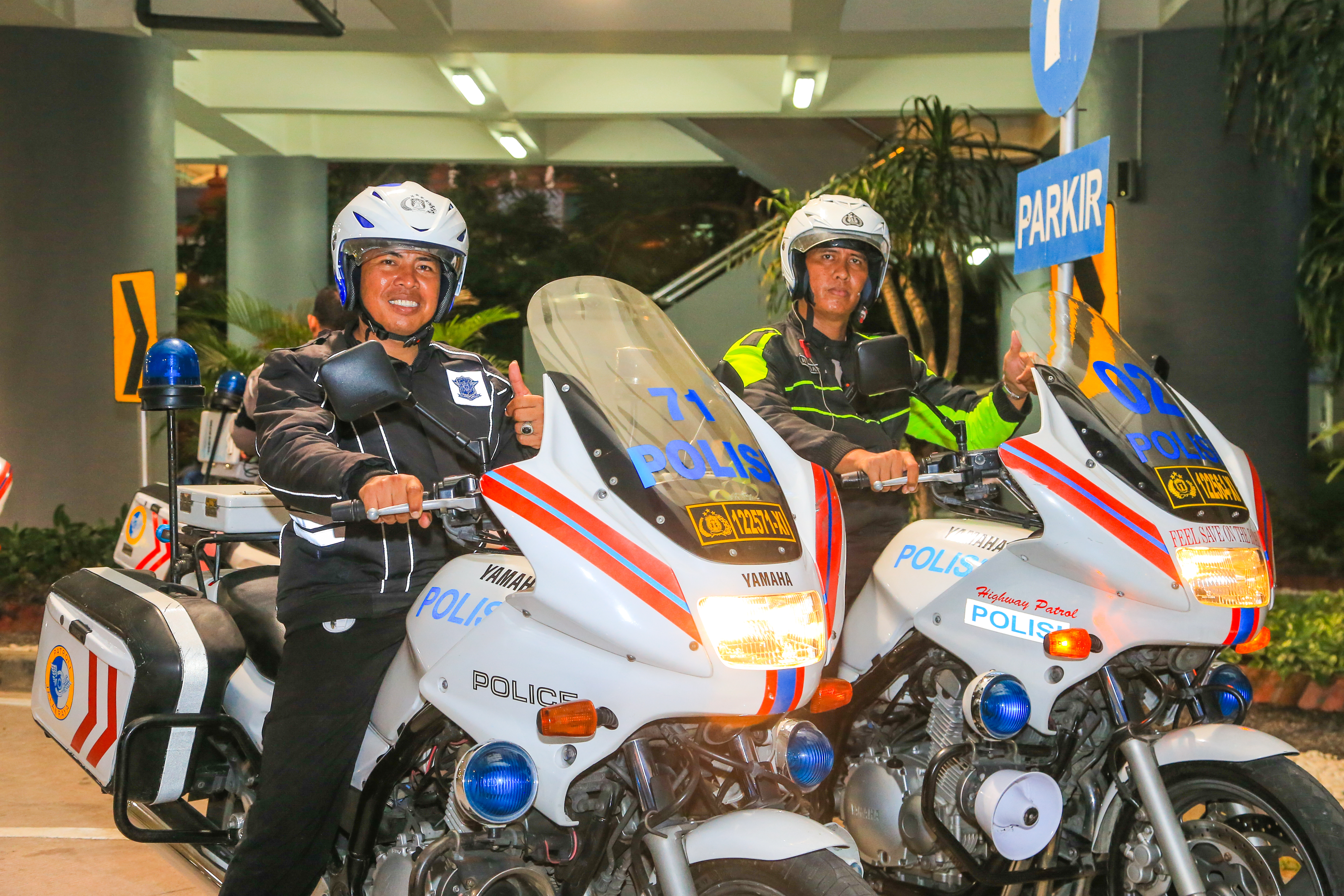 两名警员驾乘印有“POLISI”标识的警用摩托，面带笑意竖起大拇指，状态饱满，周边可见“PARKIR”标识牌。本次警摩护航专为安利活动设置，充分体现活动的高规格保障水准，氛围稳妥可靠，为参与活动的伙伴提供出行安全支持。