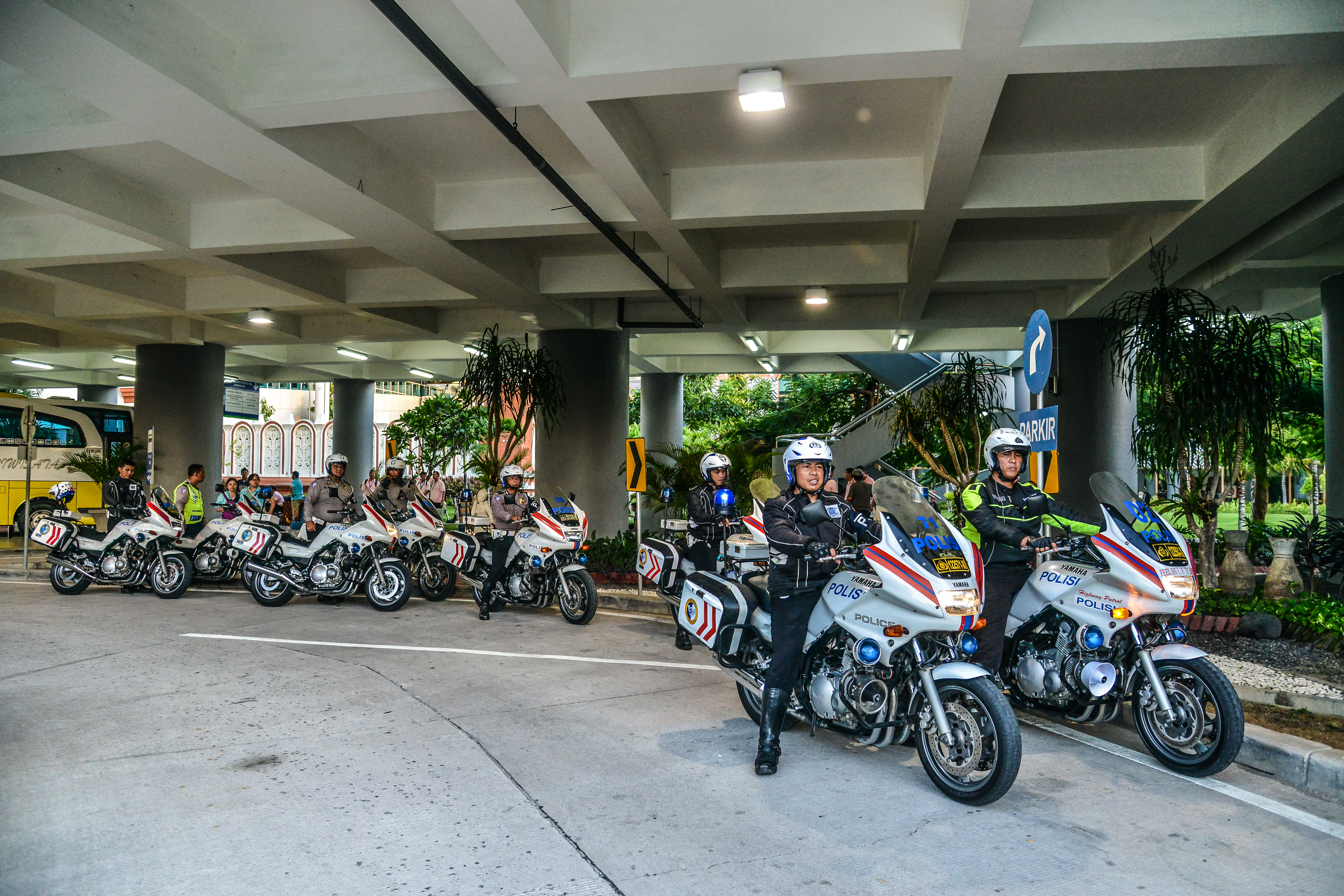画面中多名印尼警员驾乘印有“POLISI”字样的警用摩托，整齐列队于场地高架下方，周边可见“PARKIR”标识牌，警员们精神抖擞、队列规整，为本次安利海外品牌活动提供坚实安全保障，现场秩序井然，尽显活动高规格保障标准。