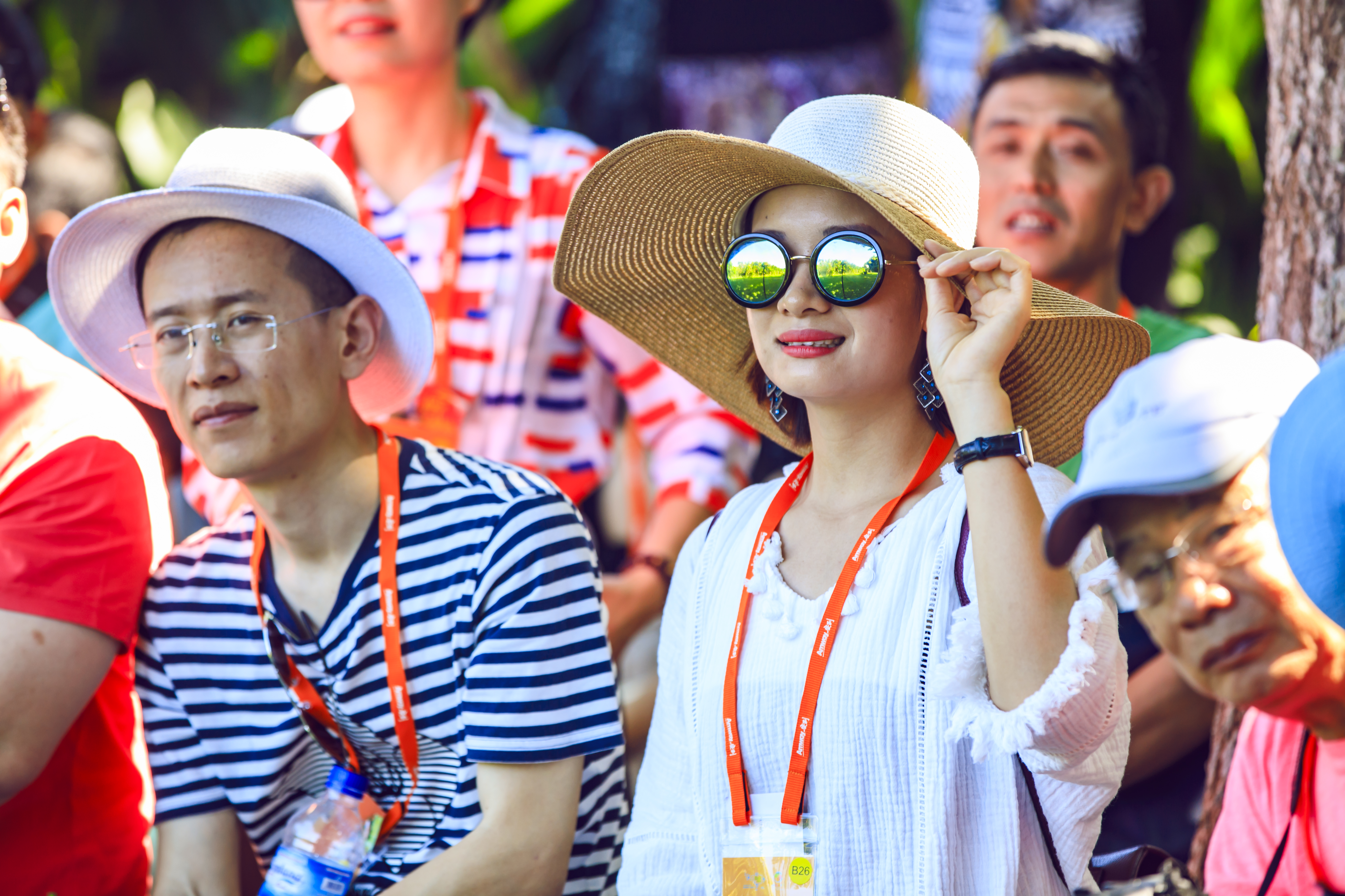 活动现场绿意环绕，参与的安利伙伴们均佩戴印有“Amway”字样的橙色专属挂绳，一位女士戴着宽檐草帽与圆框墨镜，抬手轻扶帽檐，众人神情轻松愉悦，正专注参与活动，氛围明快活力，尽显伙伴们积极向上的精神风貌。