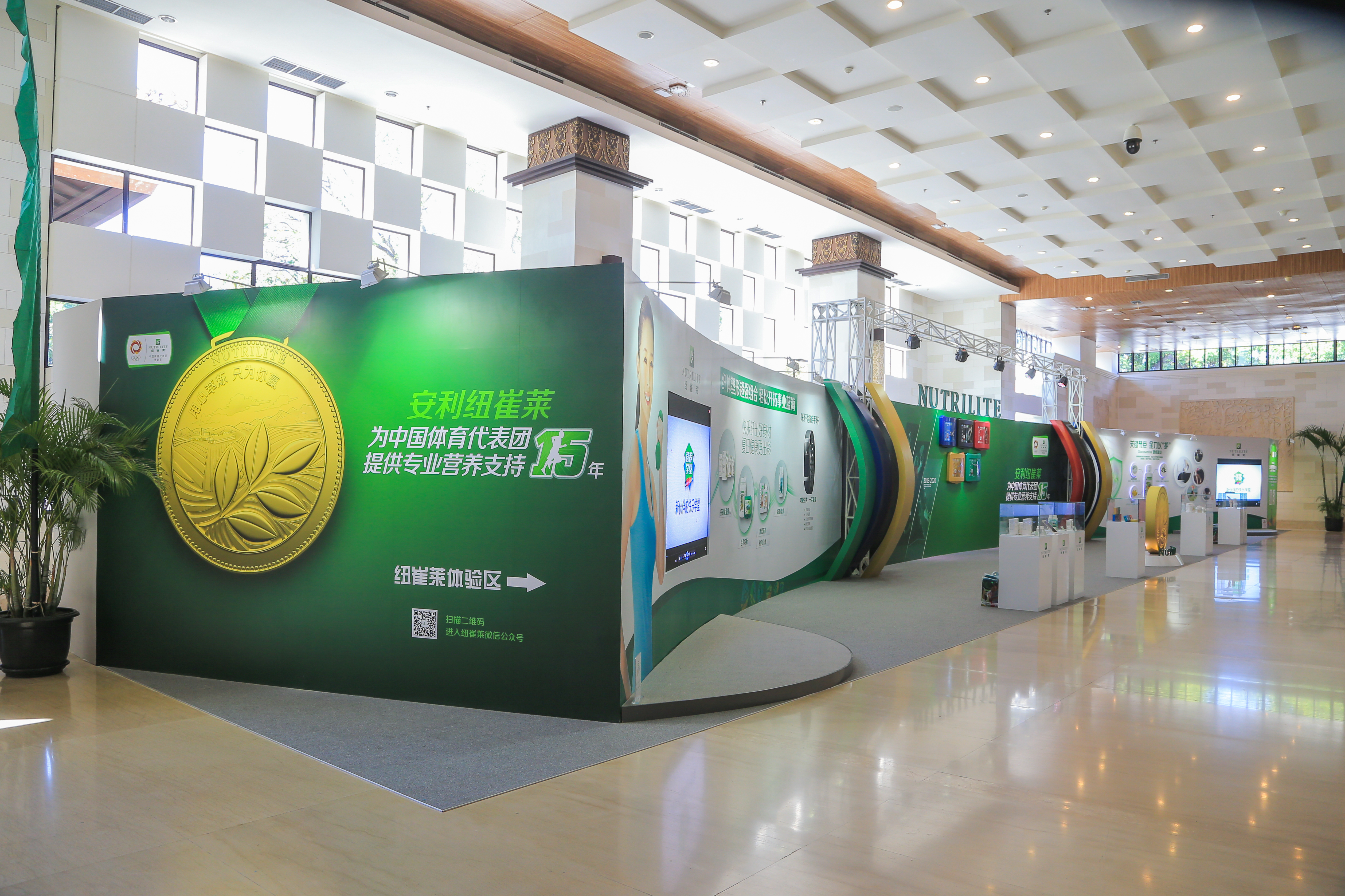 活动展厅内的安利纽崔莱展示区整洁明亮，主背景标注“安利纽崔莱为中国体育代表团提供专业营养支持15年”“纽崔莱体验区”字样，搭配金色奖牌视觉、产品展陈与导视元素，整体风格大气专业，尽显品牌正向调性。