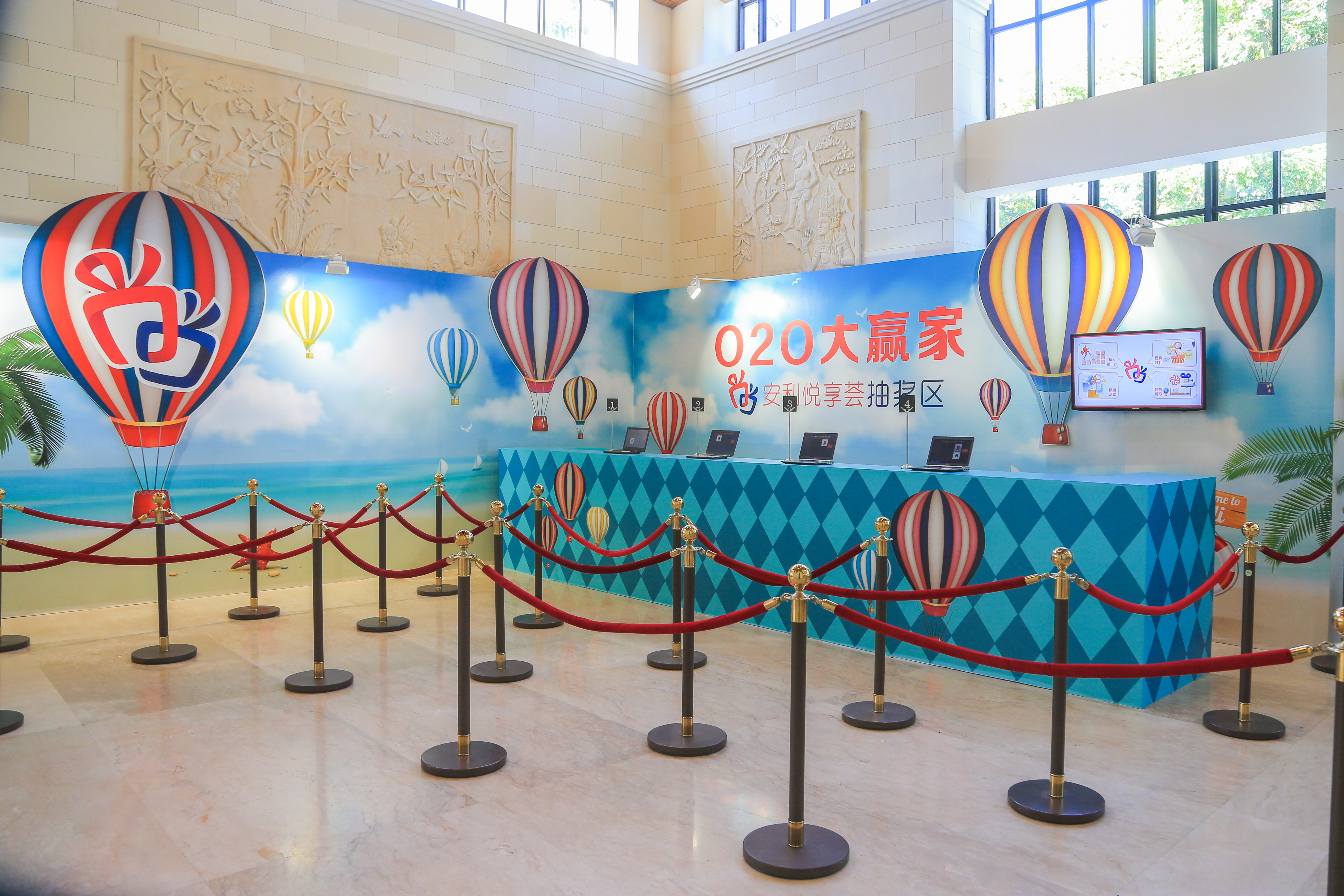 活动抽奖区以滨海热气球为主题背景，醒目展示“O2O大赢家 安利悦享荟抽奖区”字样，蓝菱格纹服务台配备多台操作电脑，现场用红绒隔离栏规划出有序等候通道，整体布置明快规整，为参与活动的伙伴提供顺畅的抽奖服务。