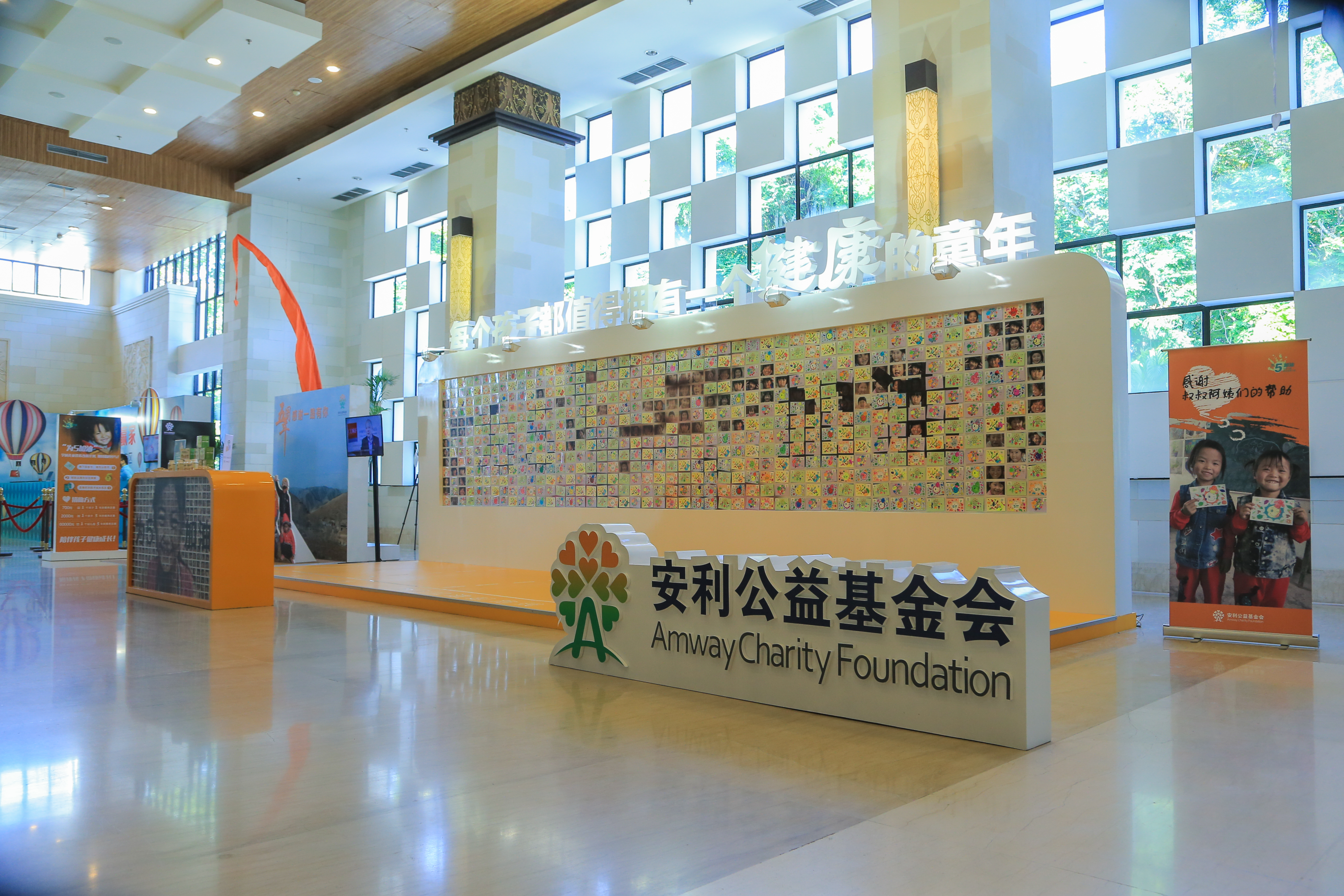 现场大厅明亮开阔，前方立有醒目的“安利公益基金会”标识牌，后方展示墙标注标语“每个孩子都值得拥有一个健康的童年”，墙身布满儿童画作与照片，旁侧设儿童公益主题展架，整体氛围温暖正向，彰显安利关爱儿童成长的公益理念。