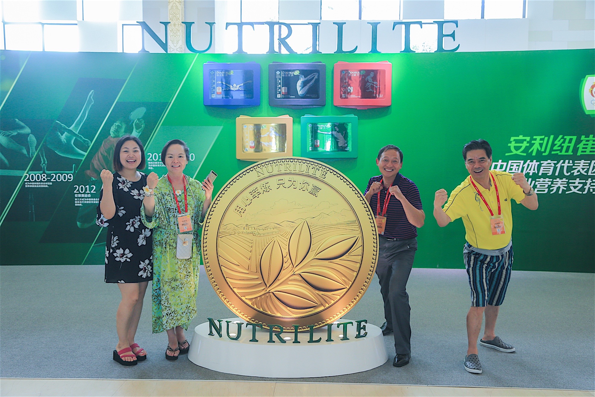 现场背景印有“NUTRILITE”“安利纽崔莱 中国体育代表团营养支持”字样，四位佩戴参会证件的伙伴站在刻有“用心萃炼 只为你赢”的纽崔莱巨型金币装置旁，面带笑容比出加油手势，氛围轻松愉悦，尽显大家对品牌的认可与参与活动的高涨热情。