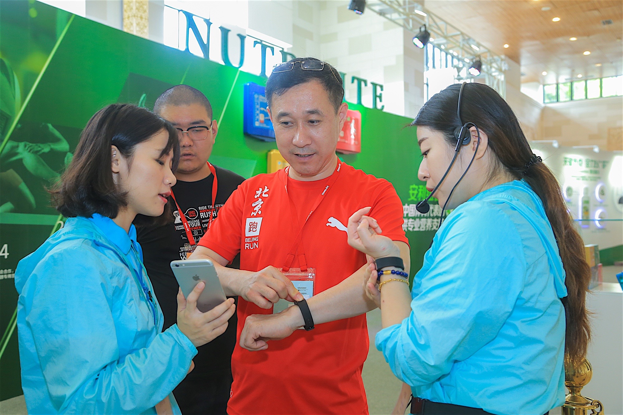 活动现场背景为印有纽崔莱、“中国体育代表团专业营养支持”字样的品牌展板，工作人员正为身着印有“北京跑”标识T恤的参与者调试智能检测手环、核对手机端数据，现场氛围专业贴心，充分展现安利深耕健康服务的品牌理念。