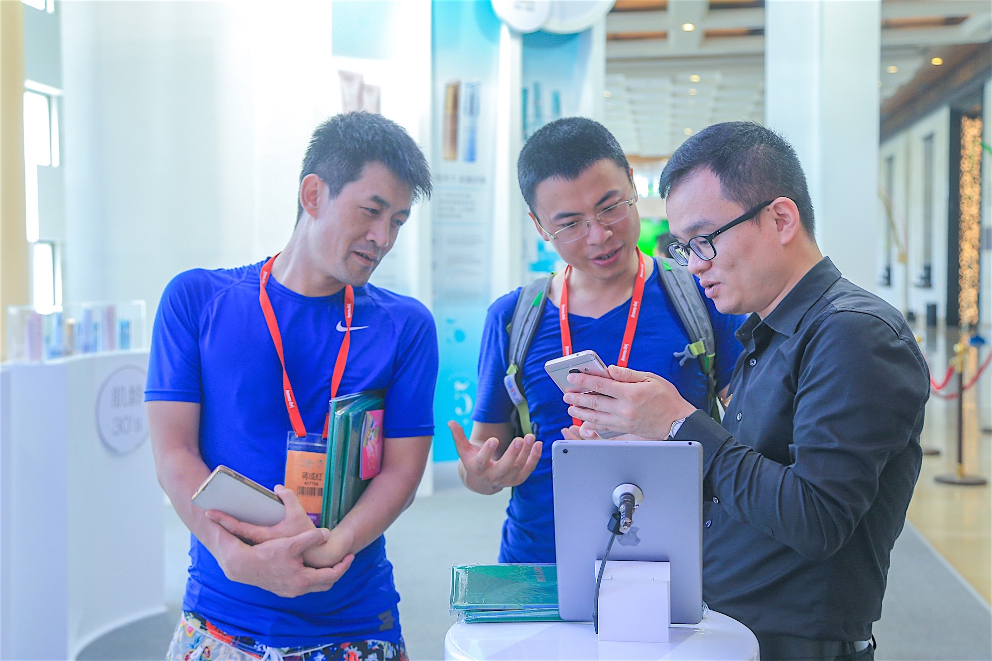 安利活动现场，两名佩戴参会挂绳的伙伴正专注聆听工作人员讲解，工作人员结合手机、展台处的平板为伙伴介绍相关内容，旁侧展区可见“肌龄30's”标识，现场环境明亮整洁，交流氛围融洽热切，尽显参会伙伴的学习热情与活动的良好互动氛围。