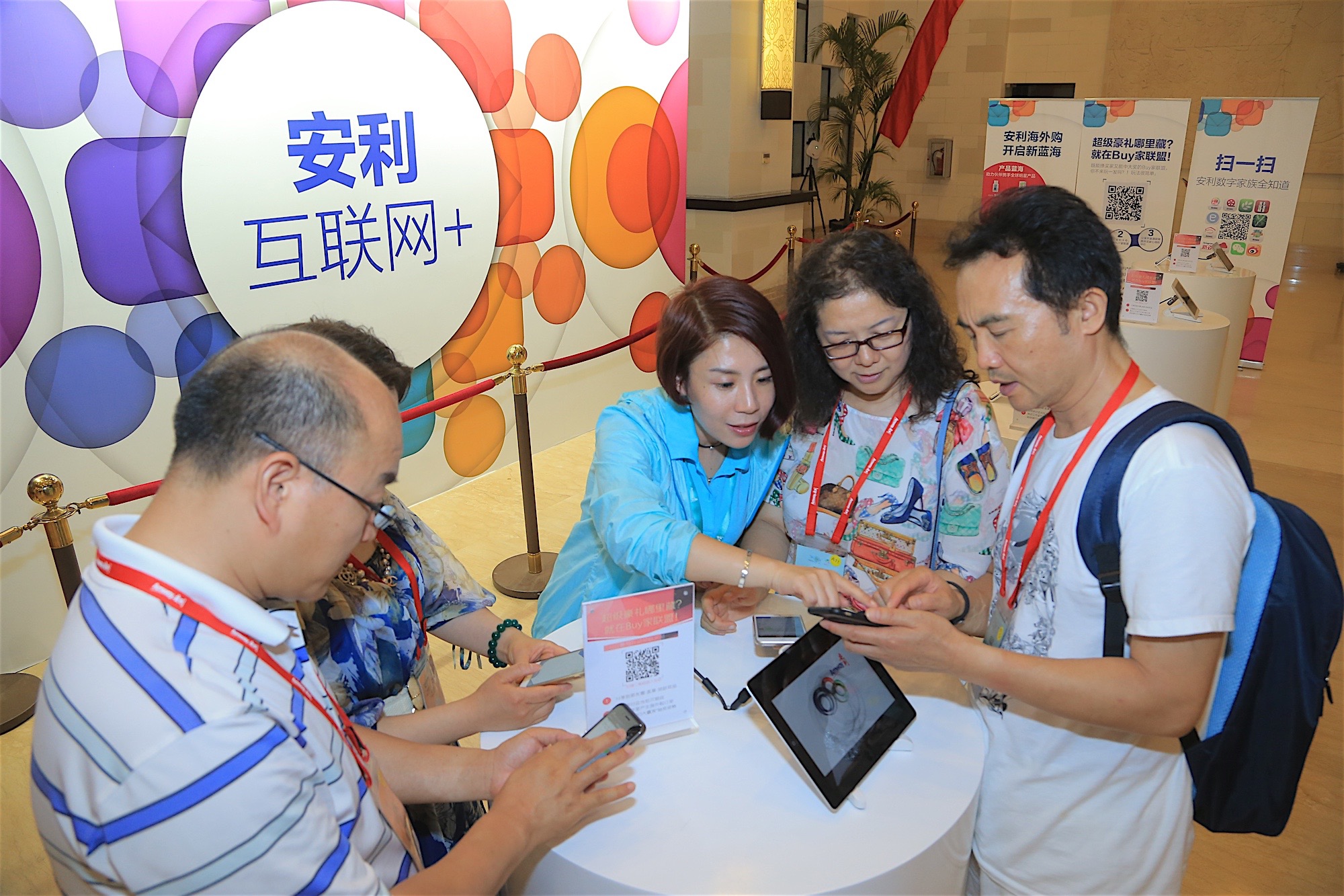活动设置“安利互联网+”主题展区，工作人员正向佩戴参会挂绳的伙伴指引操作，大家围在圆桌旁，通过手机、平板扫码参与“超级豪礼哪里藏？就在Buy家联盟！”互动活动，现场氛围活跃，充分展现安利数字化服务的便捷性与吸引力。