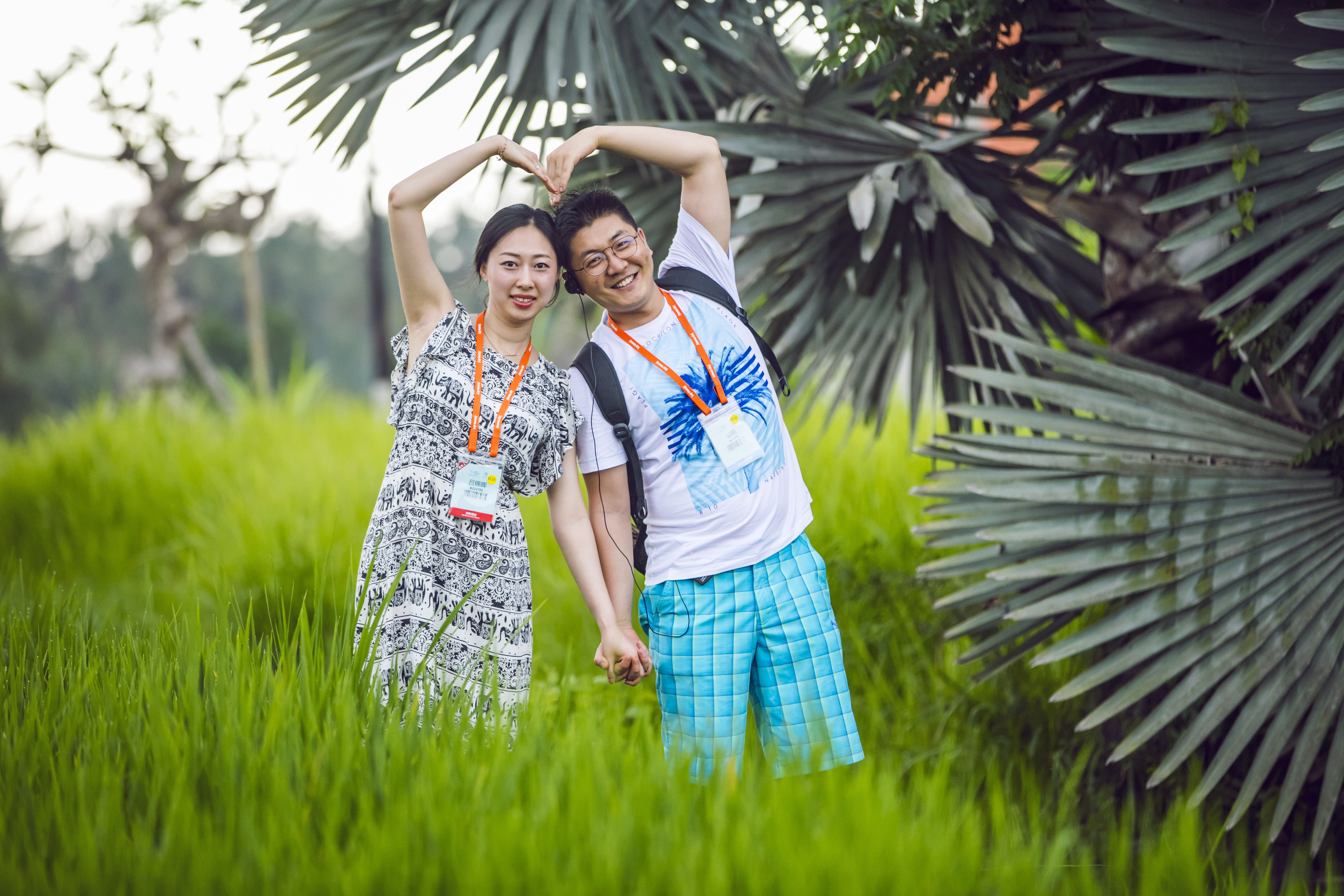两位佩戴安利活动参会证件的伙伴，伫立在鲜绿稻田与热带棕榈绿植旁，牵手举臂比出爱心造型，笑容明媚灿烂，氛围轻松愉悦，尽显安利伙伴的昂扬活力与幸福感，传递出品牌温暖正向的理念。