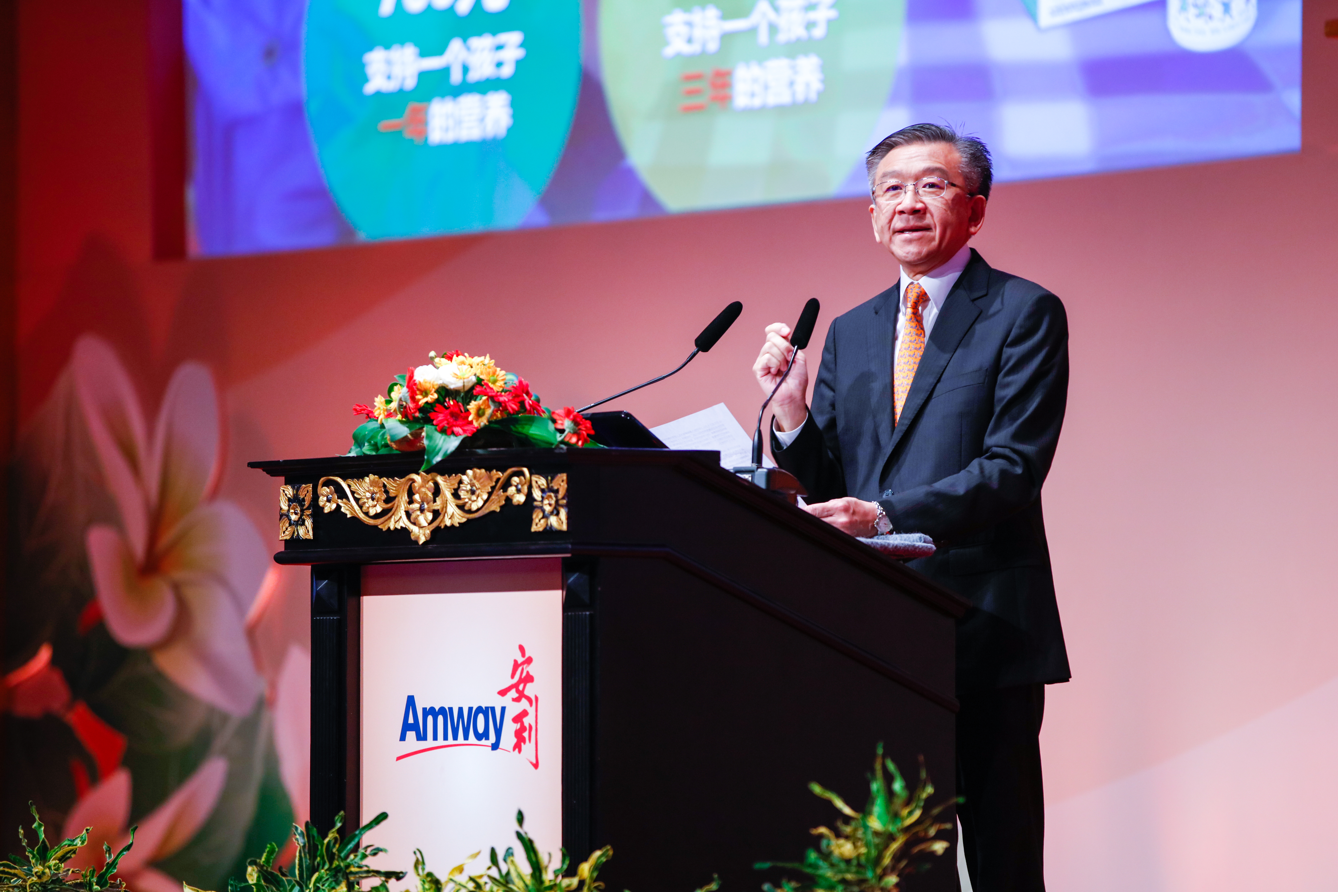 活动现场，身着正装的高管站在印有“Amway 安利”标识的雕花讲台后发言，讲台布置有鲜切花艺，背景屏幕显示“支持一个孩子 一年的营养”“支持一个孩子 三年的营养”公益相关内容，现场氛围正式积极，彰显安利践行公益责任的品牌风貌。