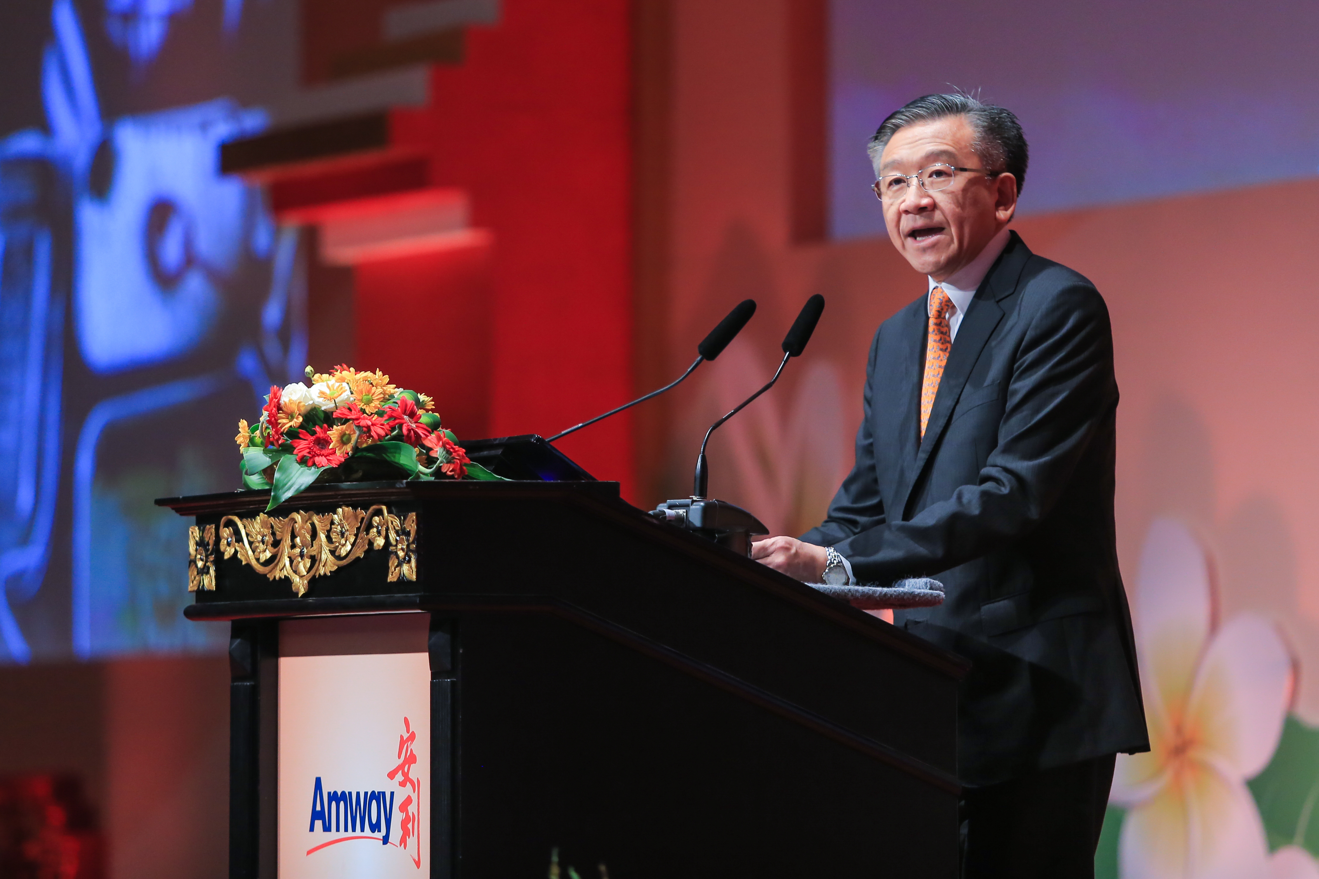 本次安利品牌活动现场，印有“Amway 安利”标识的雕花讲台上摆放着鲜切花装饰，身着正装的企业高管正站在讲台后，对着双麦面向全场发表讲话，现场氛围庄重正式，充分彰显安利积极向上的品牌风貌。