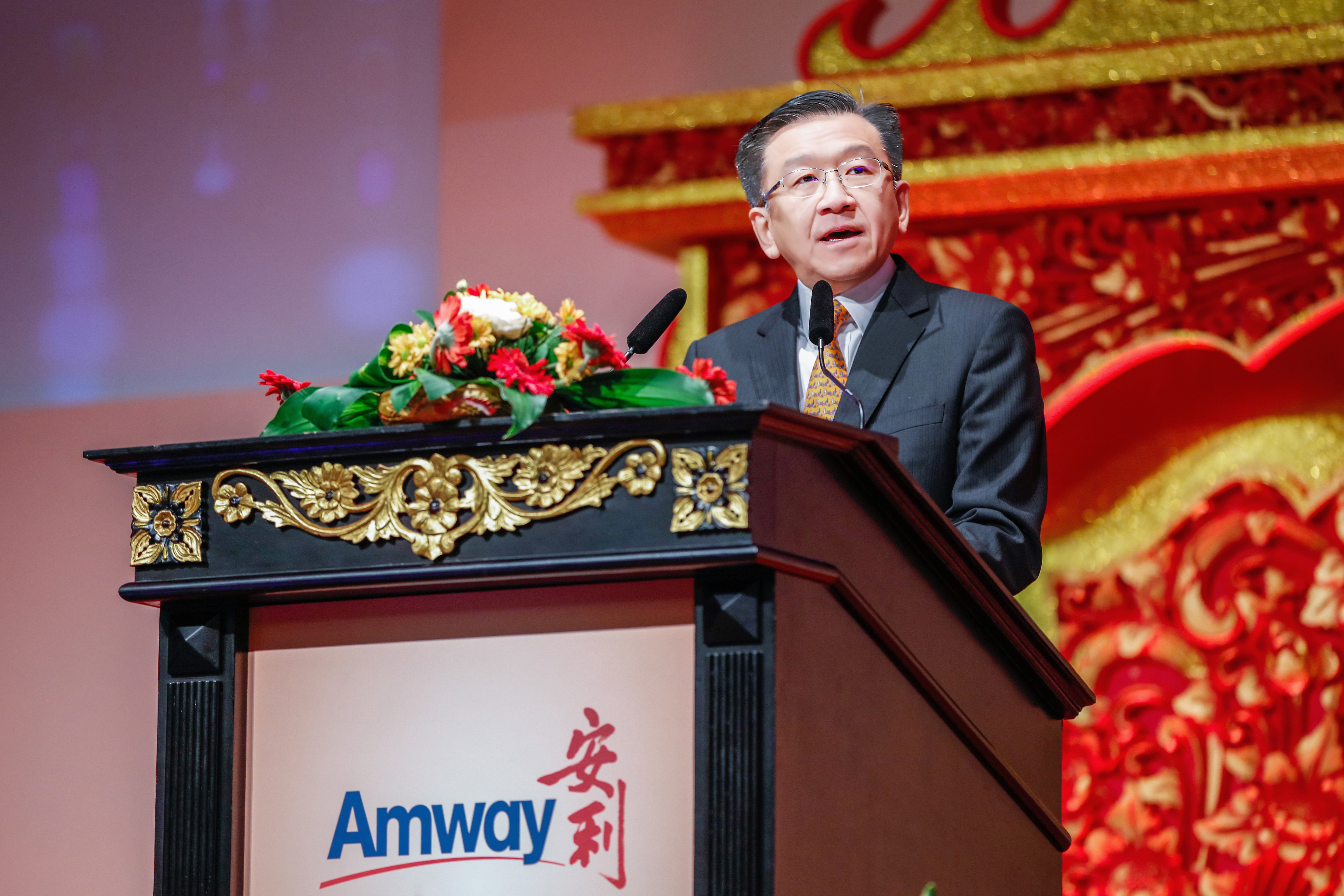 本次为安利（Amway）品牌活动现场，身着正装的企业高层站在带有精美鎏金雕花装饰的讲台后发表致辞，讲台上点缀着鲜切花艺，背景为喜庆大气的中式红金装饰，现场氛围庄重热烈，充分展现安利品牌活动的正式格调与昂扬风貌。