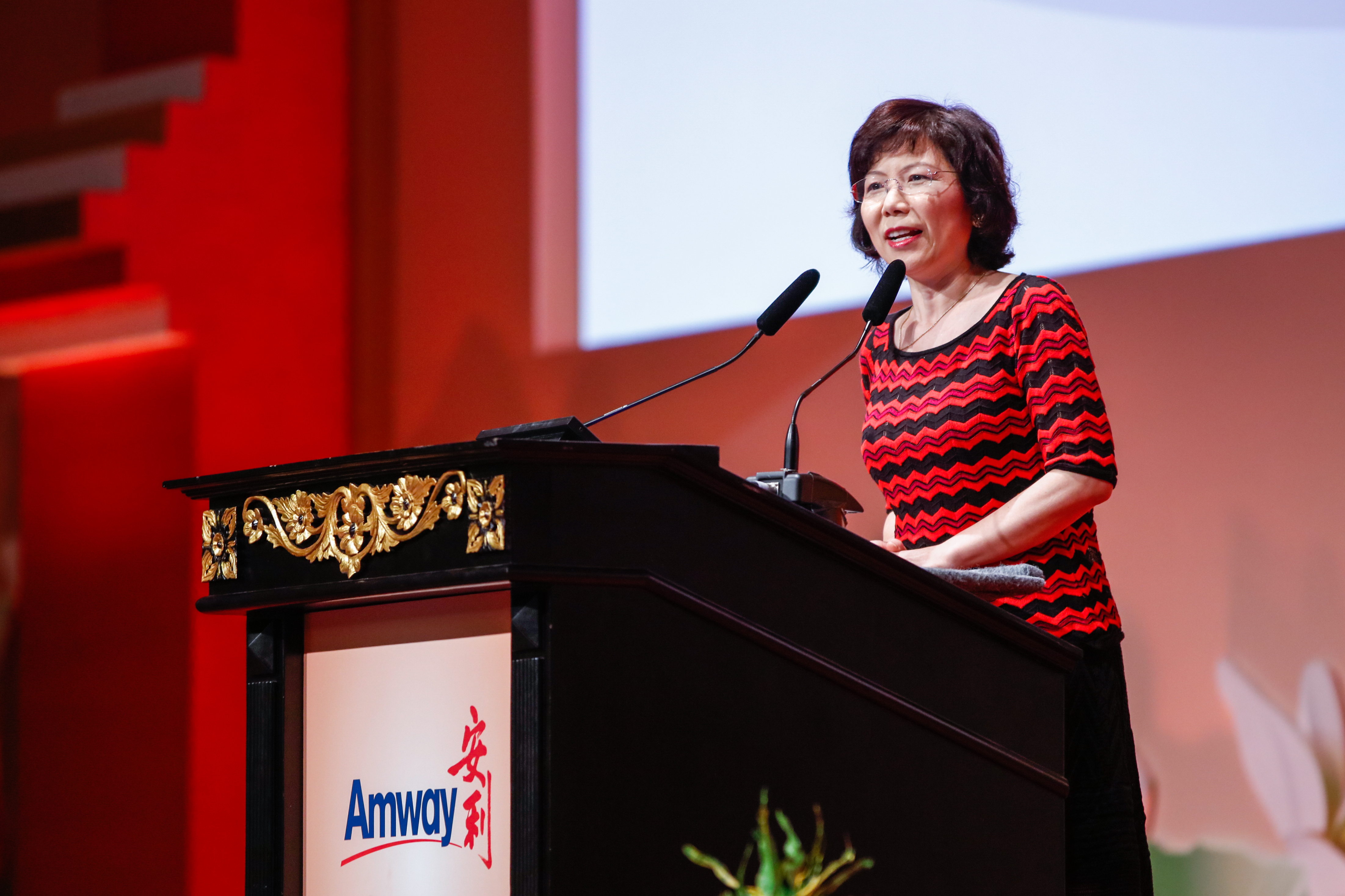 演讲台印有核心标识“Amway 安利”，一位女士站在装饰有金色雕花的演讲台后，对着话筒从容发言。舞台背景以红、白为主色调，整体氛围正式热烈，充分展现安利品牌活动的专业积极风貌。