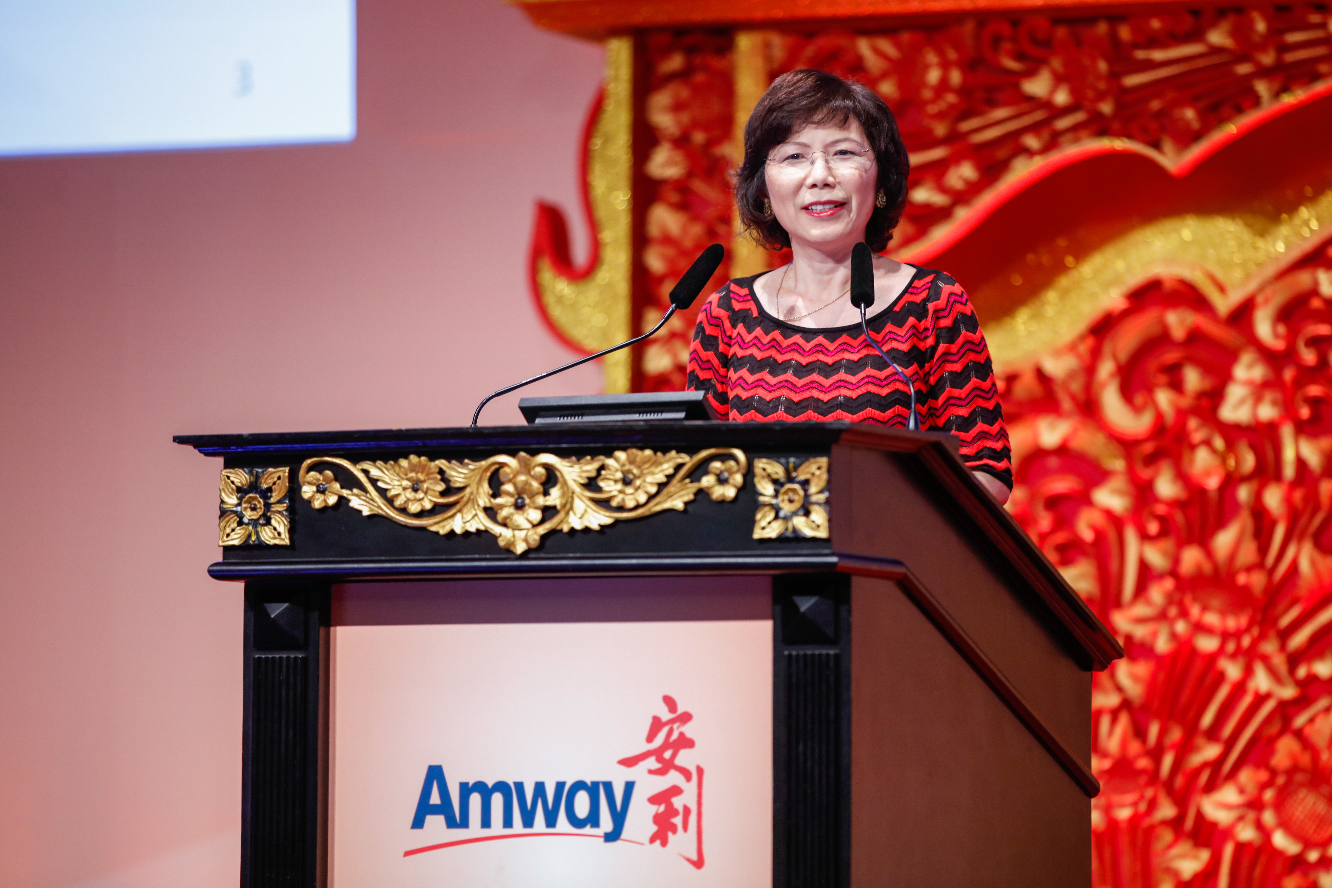 活动现场，印有“Amway 安利”标识的雕金演讲台庄重精致，背景为红金雕花中式装饰，大气喜庆。女士站在演讲台后从容发言，神态亲和，现场氛围正式热烈，尽显安利品牌活动的积极风貌。