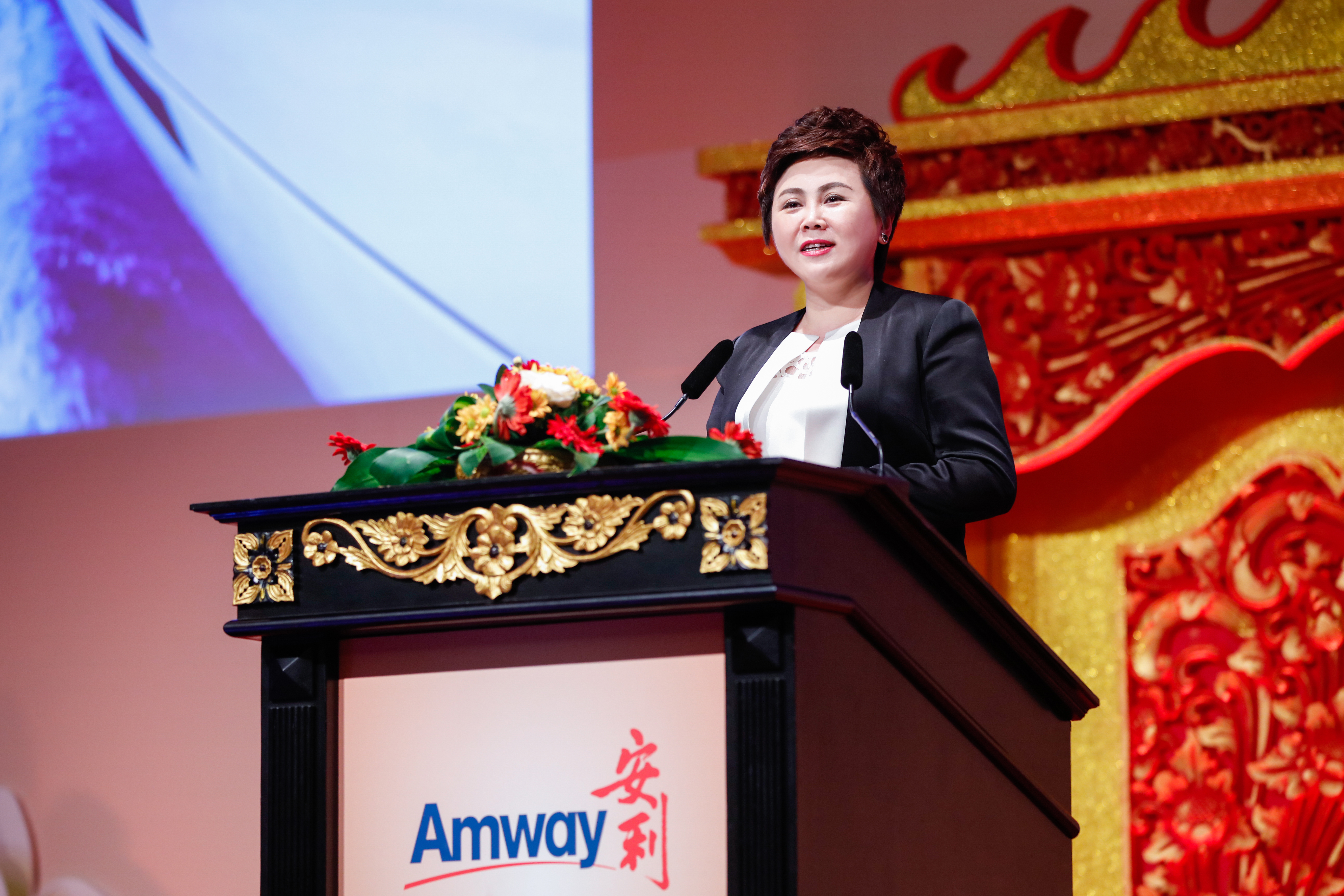 活动现场，身着正装的女性嘉宾立于印有“Amway 安利”标识的雕花讲台后发言，讲台摆放着色彩鲜亮的花艺装饰，背景搭配中式鎏金纹饰与显示大屏，现场氛围庄重热烈，尽显安利活动的专业质感与积极风貌。