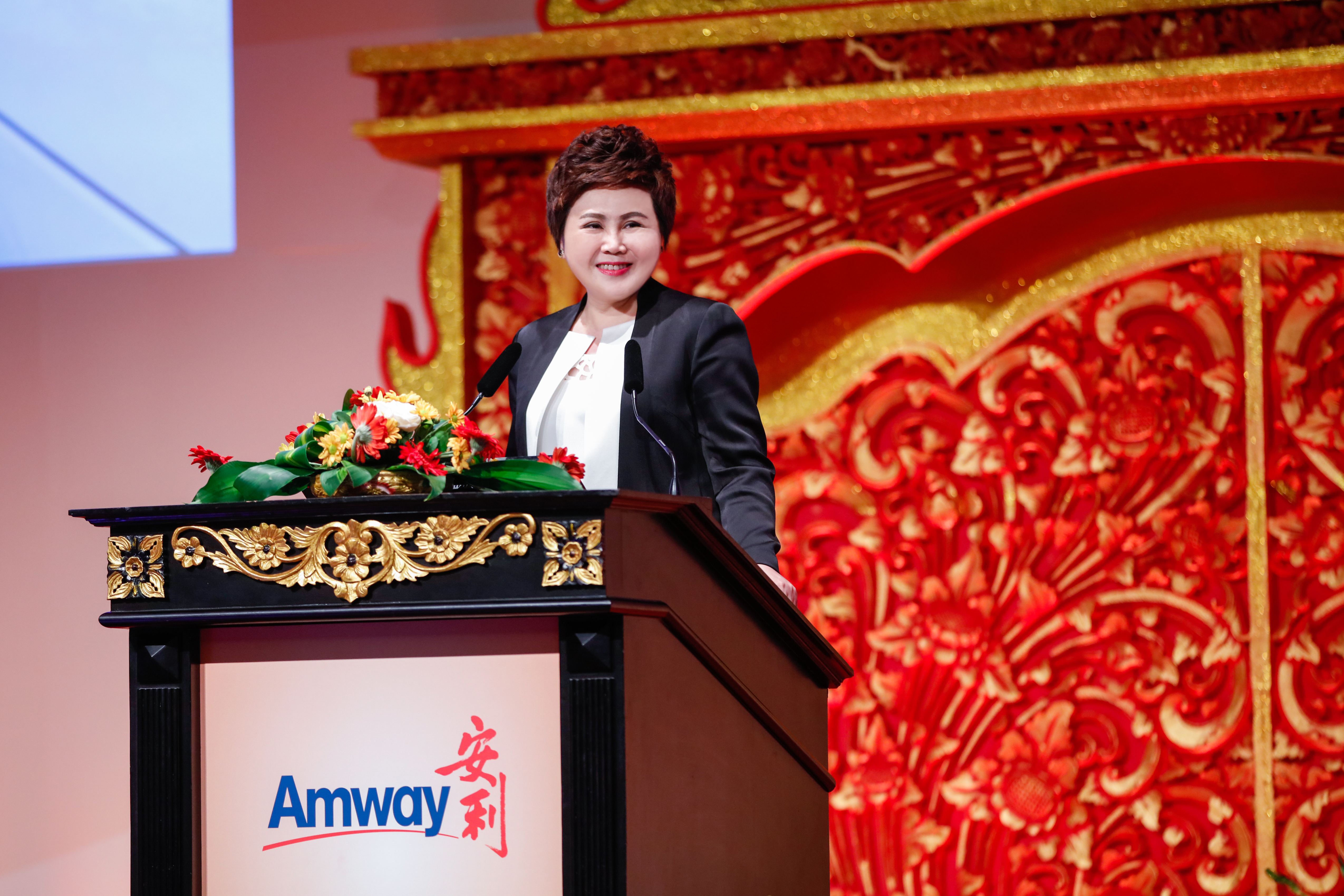 画面为安利（Amway）品牌活动现场，身着正装的女士站在饰有鎏金纹样、摆放鲜花的讲台后致辞，讲台印有安利品牌标识，背景是红金配色的精美雕花布景，整体氛围庄重热烈，尽显企业活动的正式大气，传递出积极向上的品牌风貌。