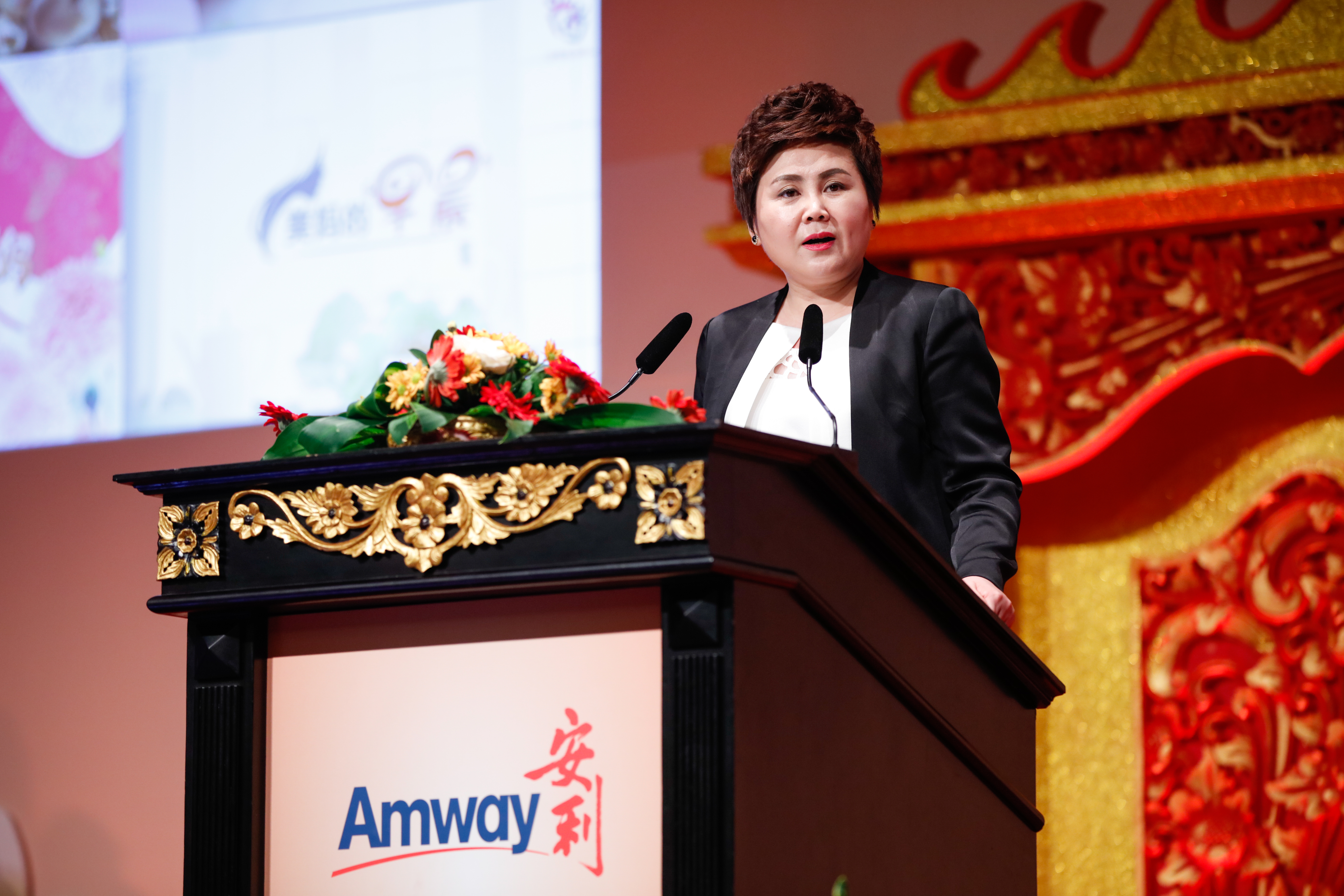 活动现场，身着正装的嘉宾立于印有“Amway安利”标识的雕花讲台后发言，讲台装点有精美鲜切花，背景搭配中式鎏金雕饰与展示大屏，现场氛围庄重正式，尽显安利品牌活动的专业风范与正向风貌。