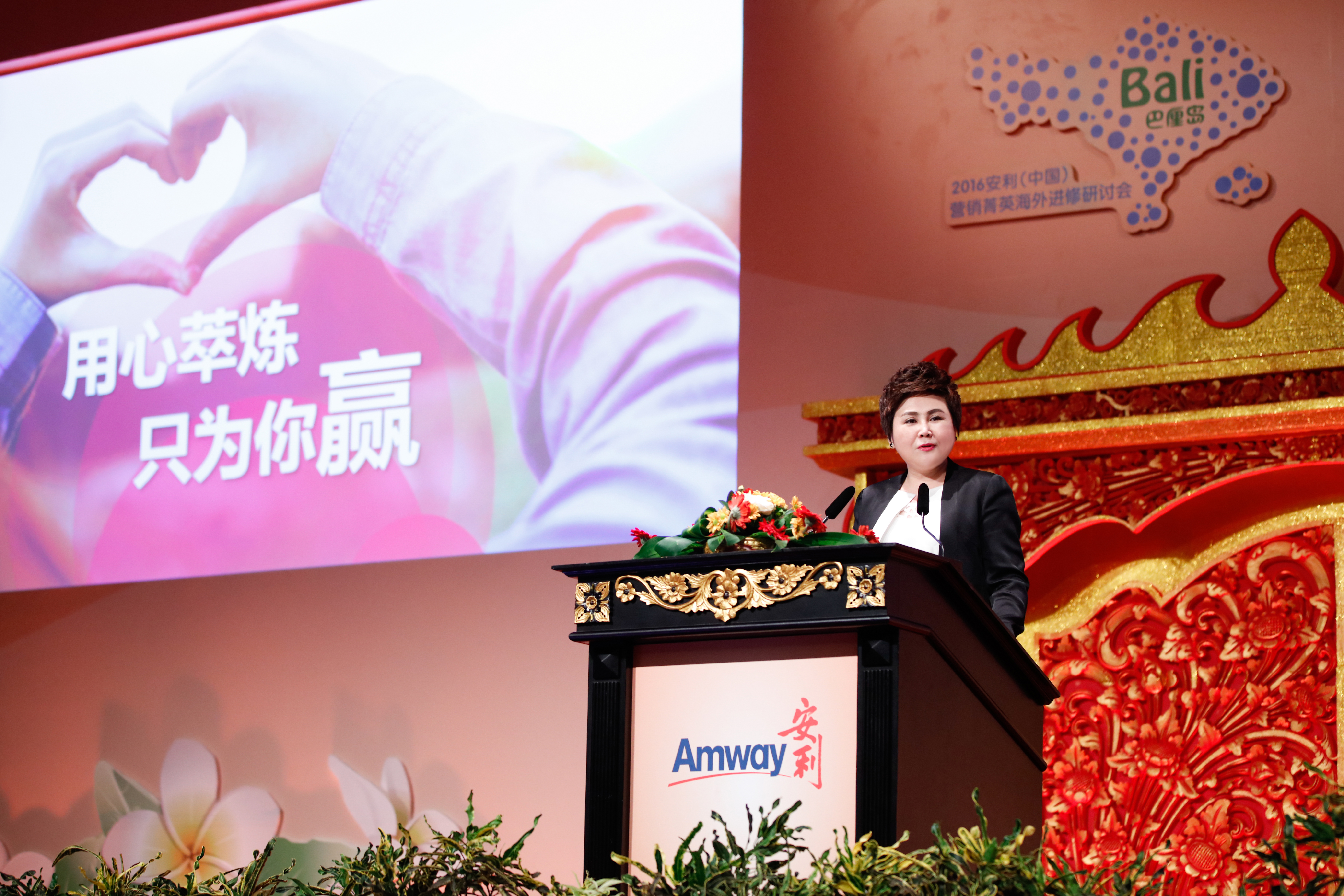 本次为2016安利（中国）营销菁英巴厘岛海外进修研讨会现场，女性嘉宾立于印有安利Amway标识的精美雕花讲台后发言，背景大屏展示“用心淬炼 只为你赢”标语与暖心比心画面，现场布置庄重精致，氛围昂扬积极，尽显品牌活动专业质感。