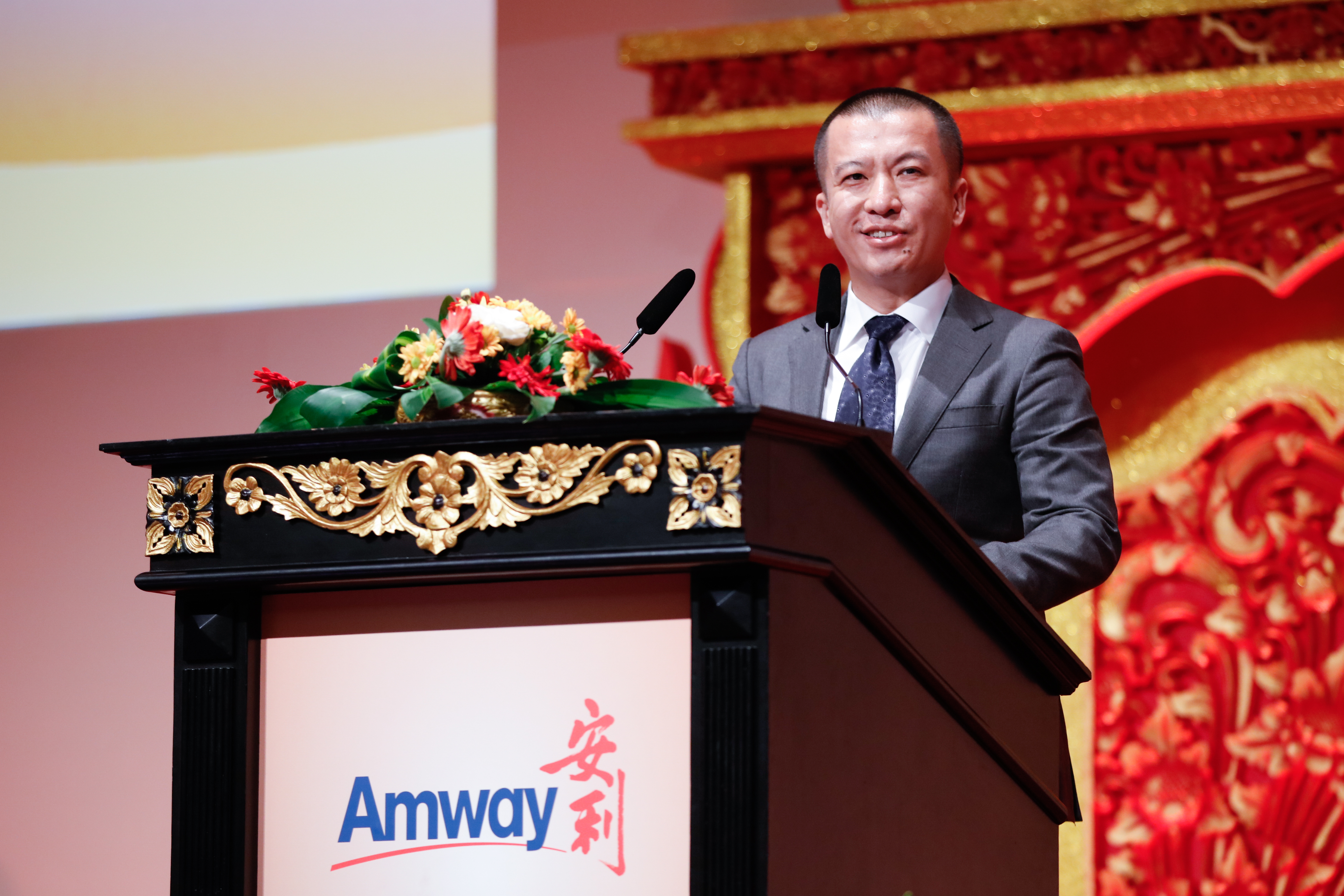 活动现场，身着正装的男士站在印有“Amway”“安利”标识的鎏金雕花讲台后发言，讲台上摆放着错落的鲜插花艺，背景为红金中式雕花装饰，整体氛围庄重热烈，尽显安利品牌活动的正式感与昂扬向上的风貌。