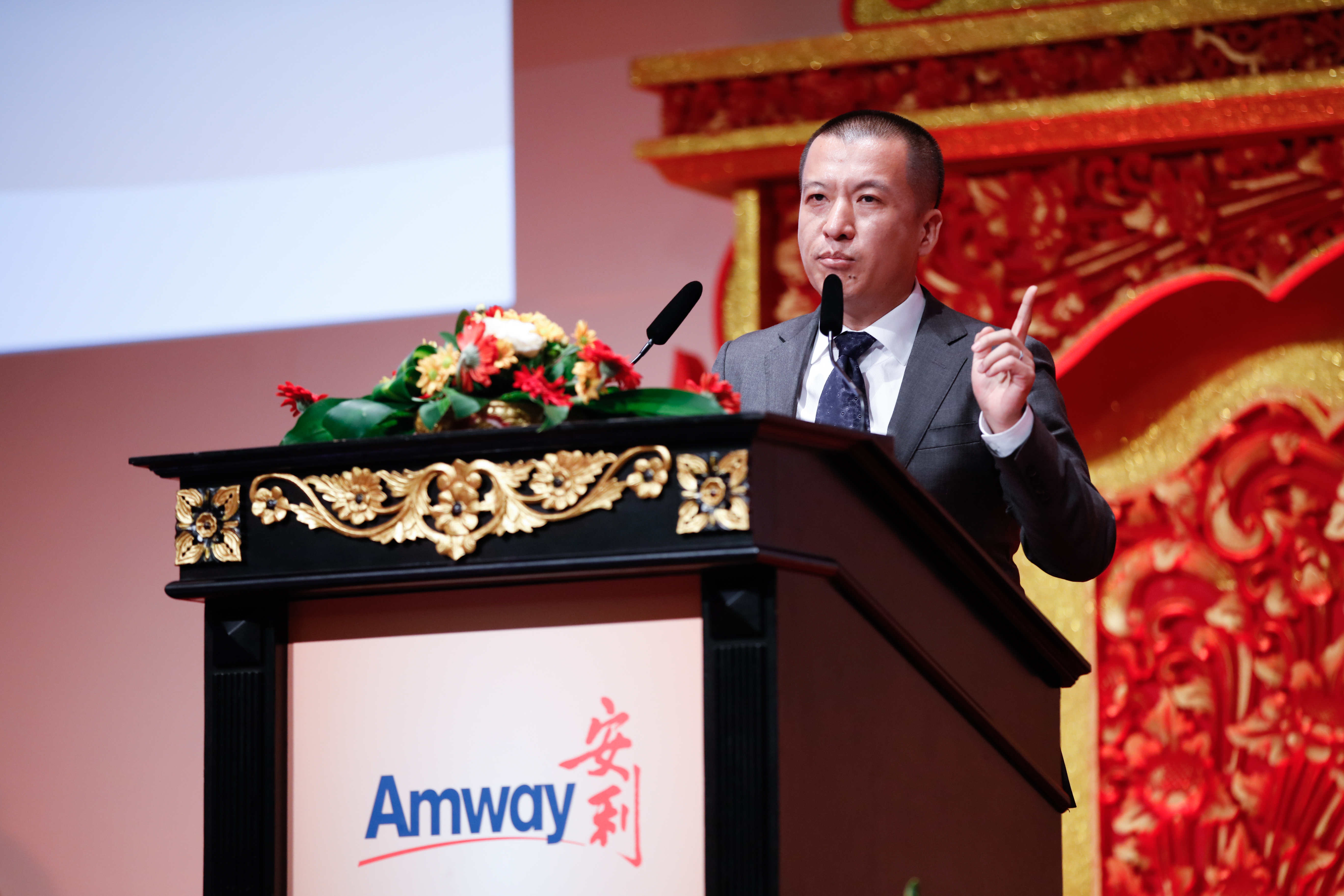 本次安利活动现场，印有“Amway”“安利”标识的精美雕花讲台上摆放着鲜花装饰，身着正装的企业高管站在讲台后抬手发言，背景红金中式雕花设计大气喜庆，现场氛围正式庄重，充分展现安利品牌活动的专业质感与昂扬风貌。