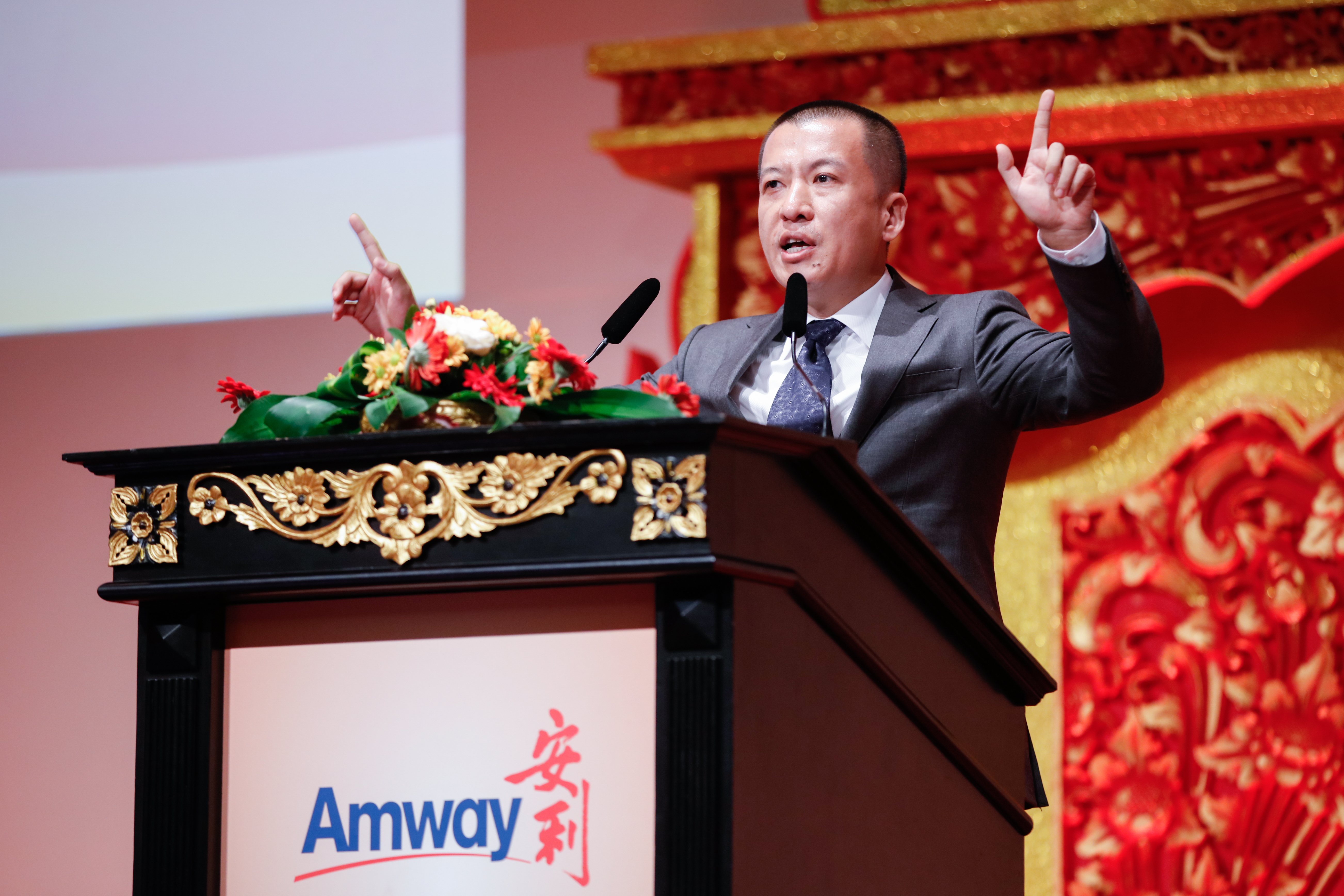 印有“Amway 安利”标识的精美雕花讲台上摆放着花艺装饰，身着正装的高管正站在台后发言，他手势有力、情绪饱满，背景红金中式雕饰庄重典雅，现场氛围昂扬热烈，尽显安利品牌活动的正式大气。