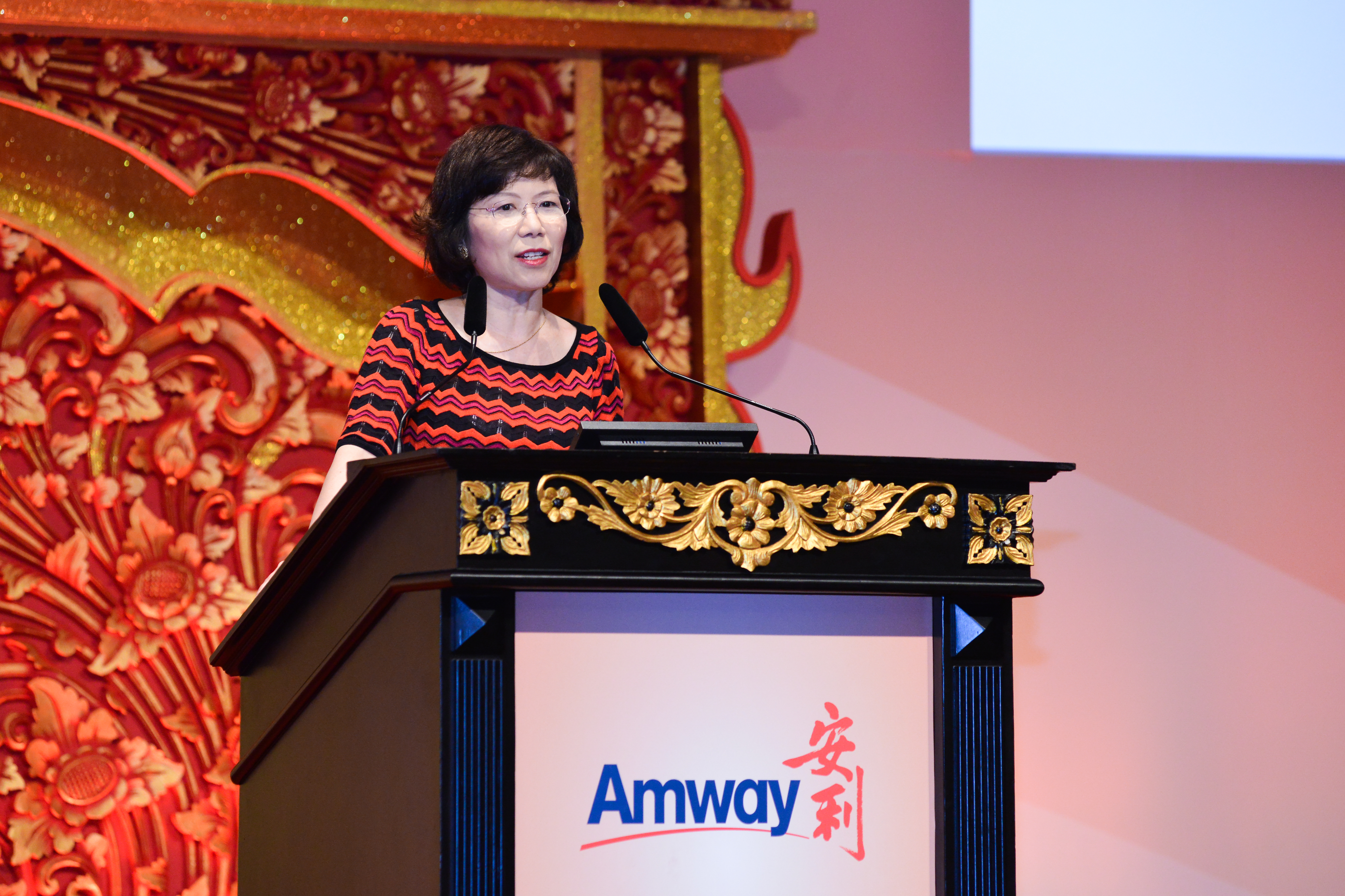 活动现场，印有“Amway 安利”标识的描金雕花讲台庄重精致，背景为红金国风雕花装饰，一位女嘉宾正站在讲台前发言，现场氛围正式隆重，充分展现安利品牌活动的专业质感与积极风貌。
