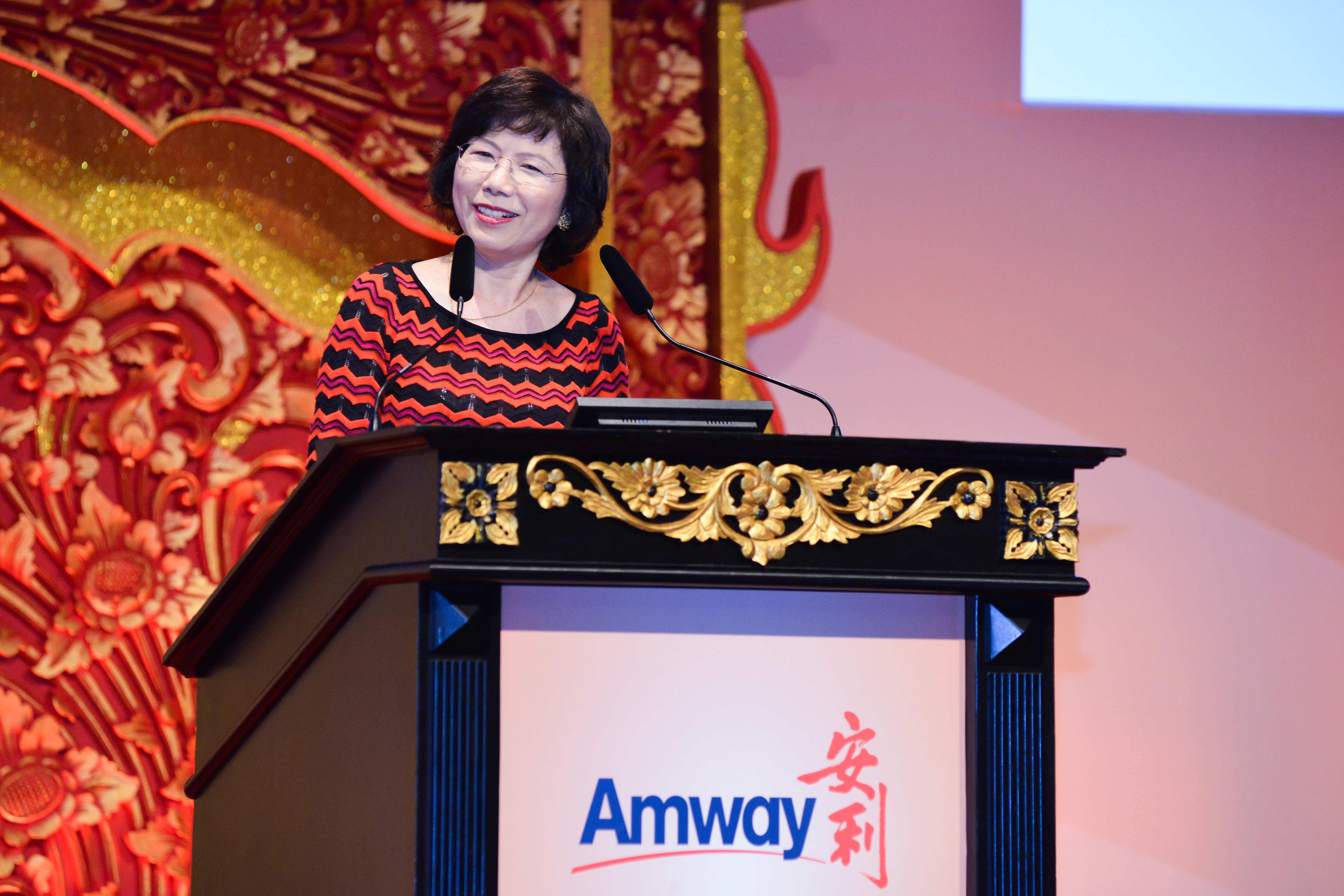 活动现场，嘉宾女士站在印有“Amway”“安利”标识的鎏金雕花讲台上发言，背景是大气的红金中式雕花布置，整体氛围庄重热烈，嘉宾神态从容亲切，充分彰显安利品牌活动专业正向的风貌。